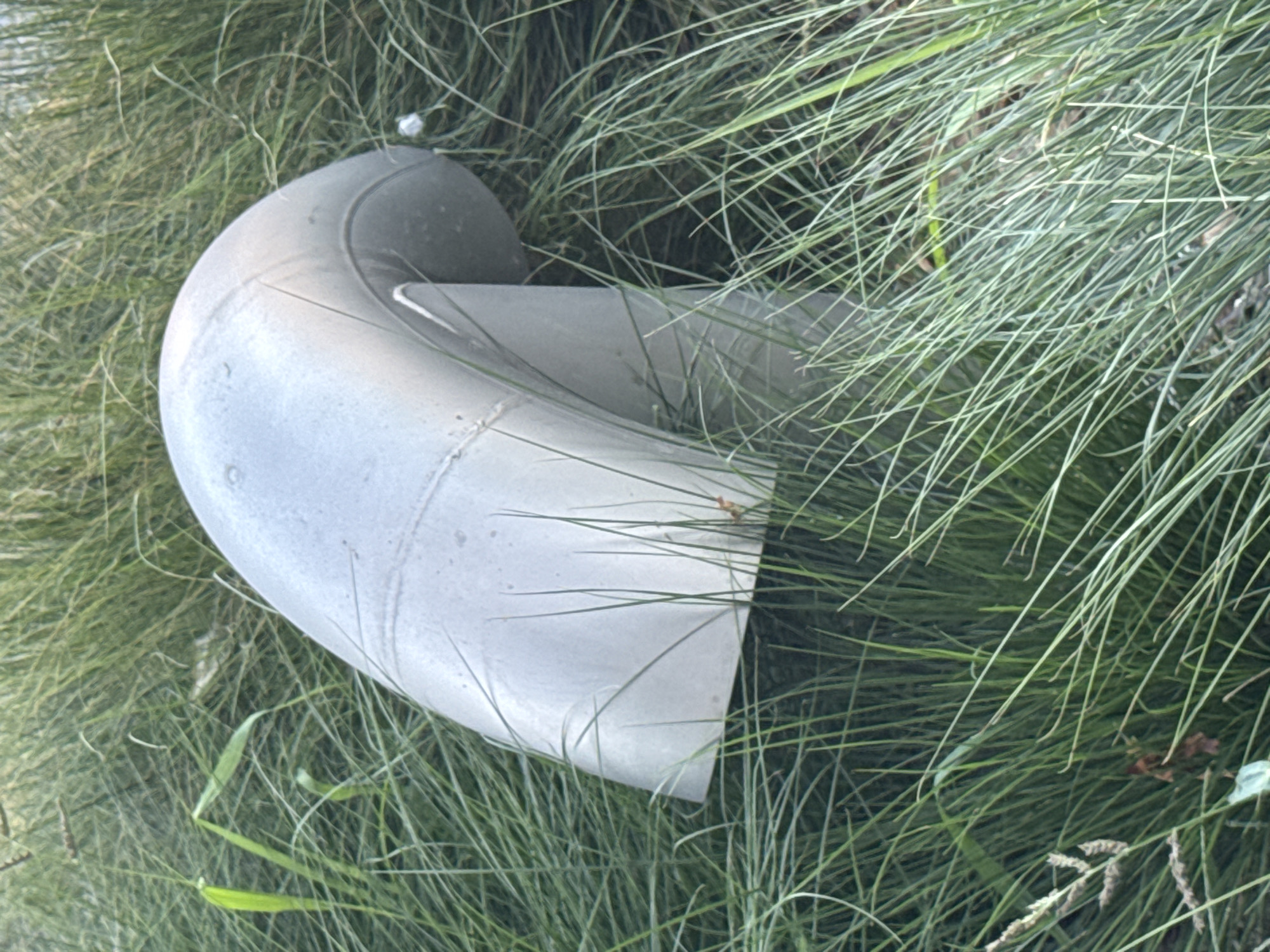 A light gray, curved pipe structure emerges from tall, dry grass in Strombeek-Bever, Belgium. The pipe has a smooth, metallic surface with visible seams. The surrounding environment is densely covered with thin, wispy blades of grass, some of which are light green and others appear dry and tan. There are no people or animals visible in the image.