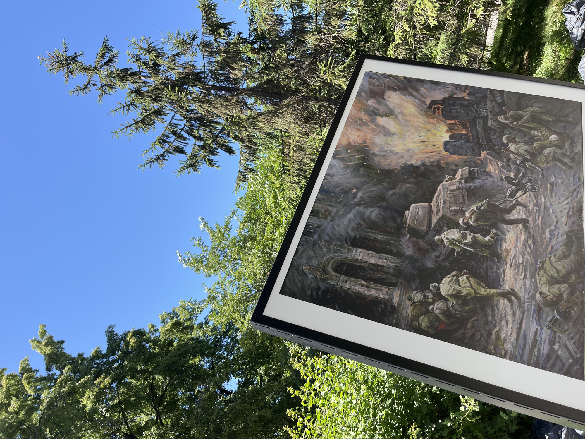 A framed painting of a military scene is displayed outdoors against a backdrop of green trees and a clear blue sky. The painting depicts soldiers in uniform advancing through a war-torn urban environment with buildings in ruins and an explosion in the background. A tank is visible on the left side of the painting. The painting is leaning against a black easel.