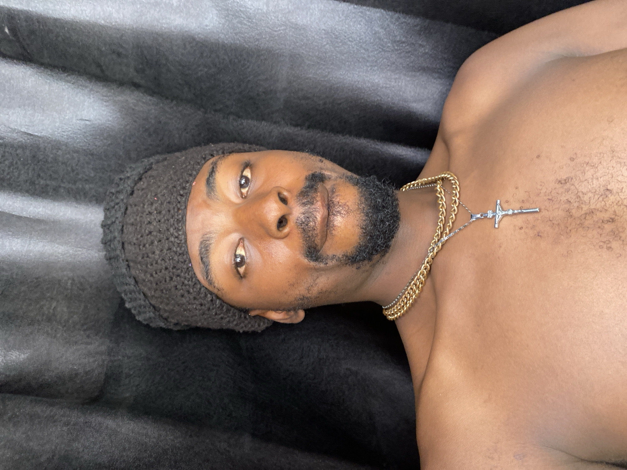 This is a medium shot portrait of a man, rotated 90 degrees clockwise. The man, who appears to be of African descent, has dark skin, a neatly trimmed beard, and dark eyes. He is wearing a dark, possibly charcoal-grey, knitted beanie that covers the top of his head. Around his neck, he sports a thick, gold-colored chain necklace layered with a thinner silver-colored chain that features a prominent silver cross pendant. He is shirtless, revealing his bare chest and shoulders, which have some scattered hair.
The man is lying down, with his head positioned on the left side of the frame and his body extending towards the right. He is looking directly into the camera with a neutral to slightly serious expression. The background consists of a dark, textured fabric, possibly a blanket or bedding, which is out of focus, creating a shallow depth of field that emphasizes the subject. The lighting is soft and even, without harsh shadows, suggesting an indoor setting or diffused light. There is no visible text in the image. The scene could be taking place in Barakin Akawo, Nigeria.