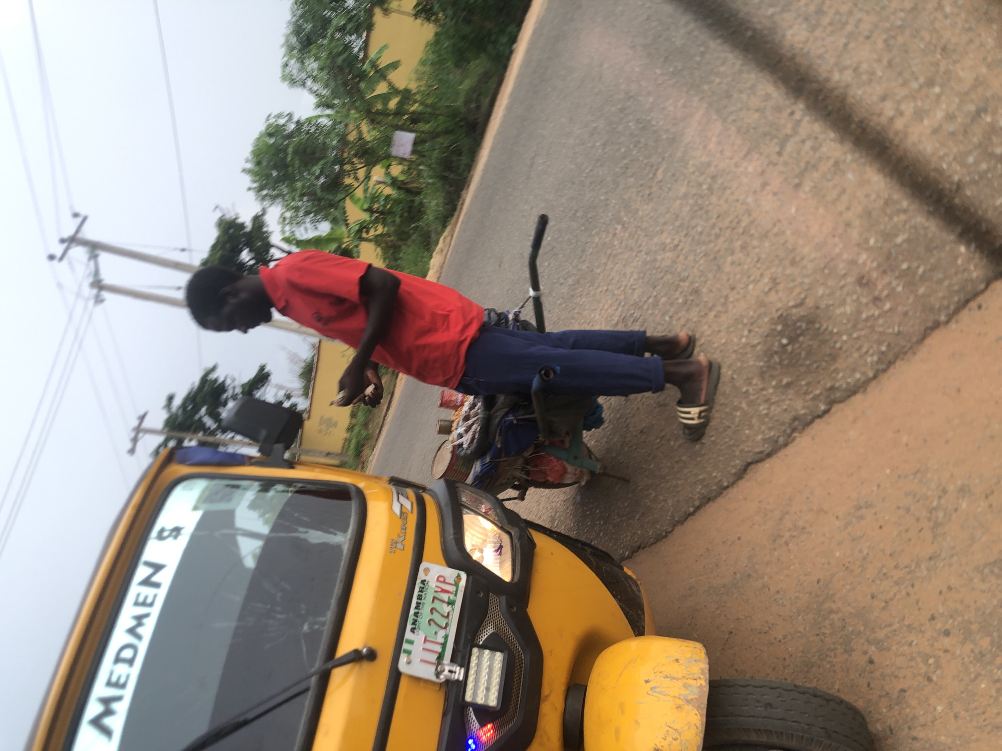 The image shows a man in a red shirt and blue pants standing next to a yellow tricycle (Keke) in what appears to be Ukwu-Abwa, Nigeria. The tricycle has "MEDMEN S" written on the front windshield and a license plate that reads "LT-227YP" with "ANAMBRA" above it. The man is looking down and appears to be holding something in his hand. The environment consists of a paved road, green foliage, and utility poles in the background.