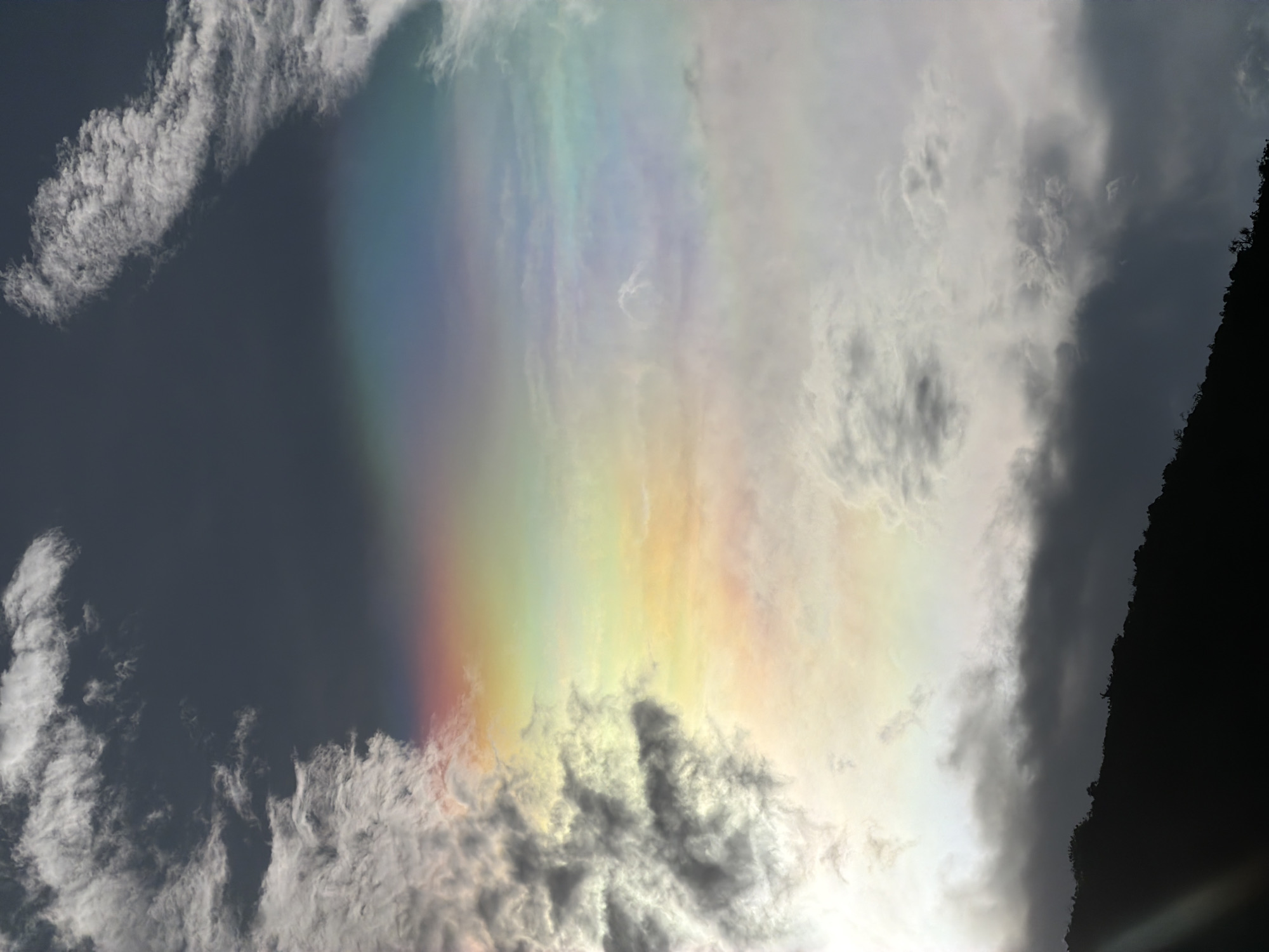The image shows a cloud iridescence phenomenon in the sky. The colorful, rainbow-like effect is visible through a break in the clouds. Other clouds are present, some darker and some lighter. The exact location in Nha Trang Ward cannot be determined from the image content. There are no people, subjects, objects, actions, or interactions visible. The environment is the sky and clouds.