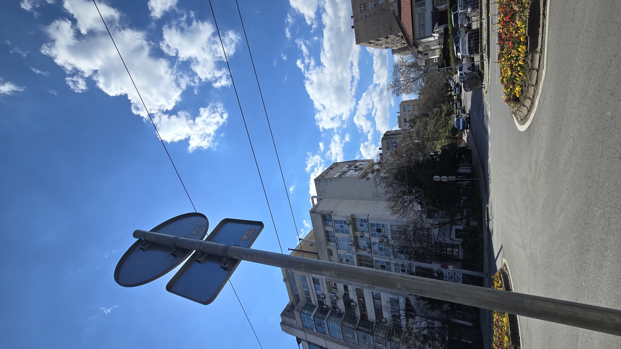 The image depicts a street scene in Stara Zagora, Bulgaria. The view is directed upwards, capturing a traffic sign in the foreground, a street, buildings, and a sky filled with clouds. The traffic sign consists of two blank blue signs attached to a gray metal pole. Power lines are visible against the blue sky with fluffy white clouds. A row of buildings lines the street, showing a mix of residential and commercial structures. Parked vehicles are also visible along the side of the road. A flower bed encircles a roundabout island next to the road.