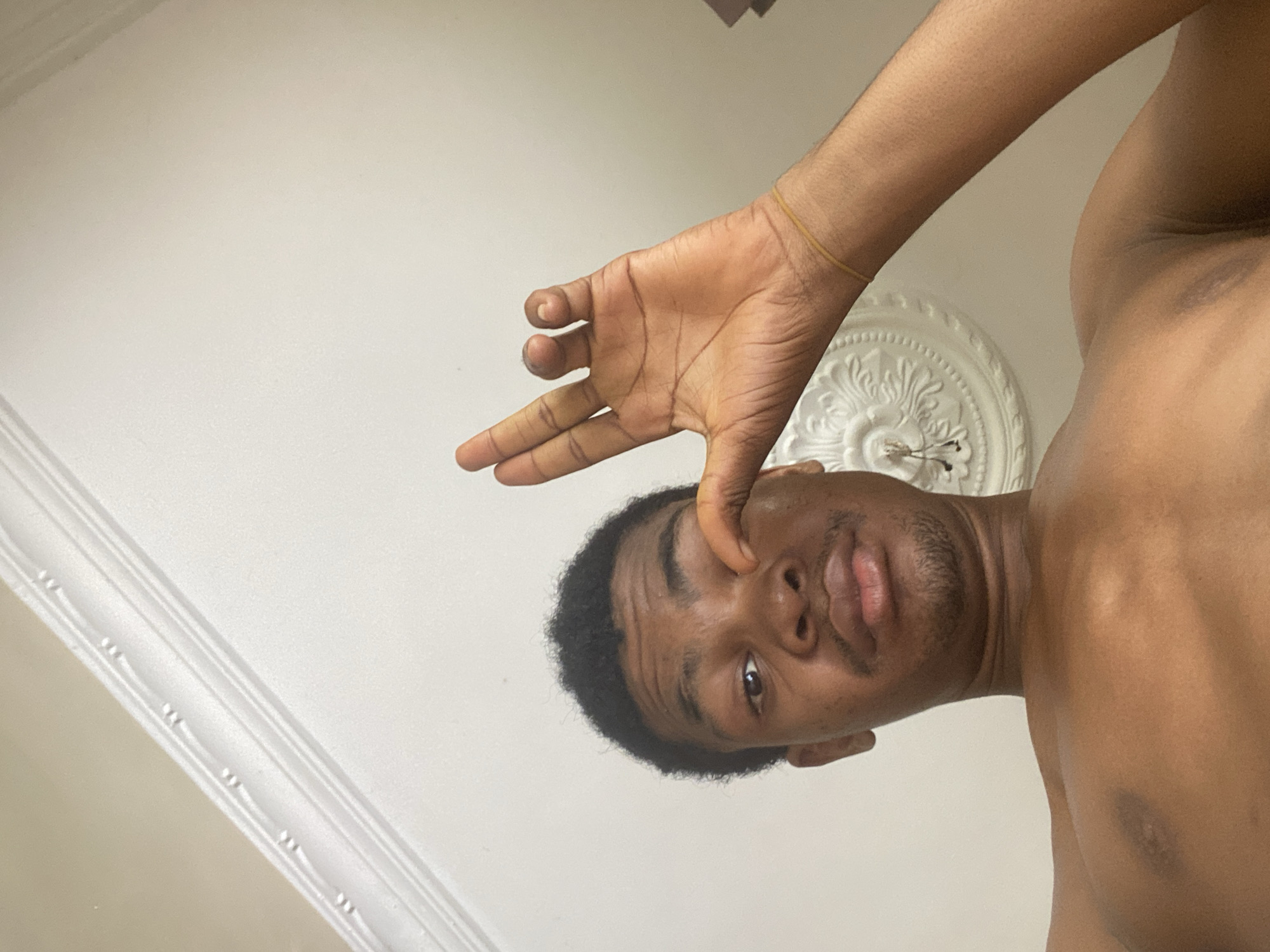 A man with dark skin, short dark hair, and a bare chest is pictured from a low angle against a white ceiling. His face is close to the camera, with one eye visible. He is making a hand gesture near his eye. The ceiling is plain except for a decorative circular element. The location is in a building, not necessarily in the city of Ado, Nigeria.