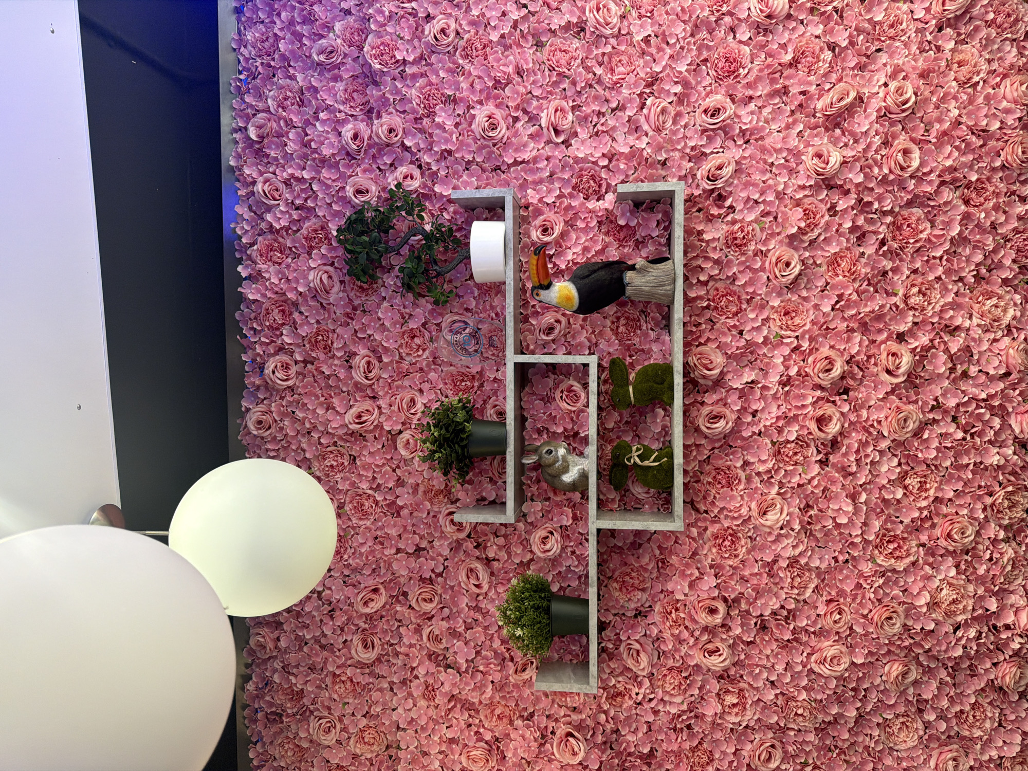 The image shows a wall covered in pink artificial flowers, with a pink and purple gradient effect. There are three light grey shelves mounted on the wall. The top shelf has a small green potted plant and a white pot with a green Bonsai tree. The middle shelf has a gray bunny figurine, and the bottom shelf has two green bunny figurines. A black and white toucan figurine is perched on the middle shelf. Two white balloons are visible in the top left of the image, hanging from the ceiling. The scene appears to be indoors, possibly a shop or a decorative display. The exact location in Greater London, United Kingdom, is not discernible from the image.