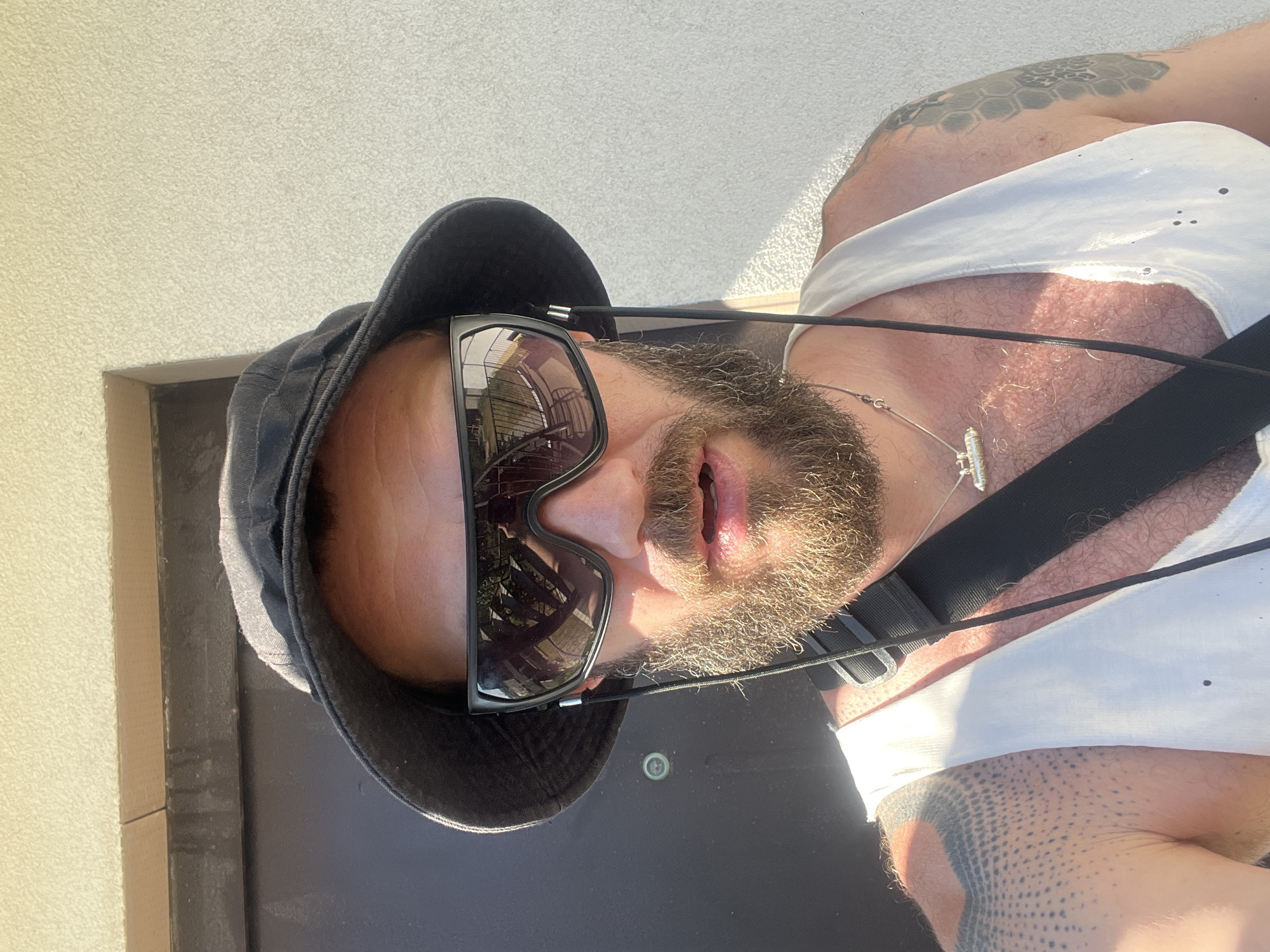 The image is a selfie taken outdoors in Балаклавский округ, Ukraine, during the daytime. A man with a beard and tattoos on his arms is wearing a black bucket hat, large black sunglasses, and a white tank top. A black strap crosses his chest. He appears to be standing in front of a building with a textured wall and a dark doorway. The sun is casting a strong light, creating shadows and highlights.
