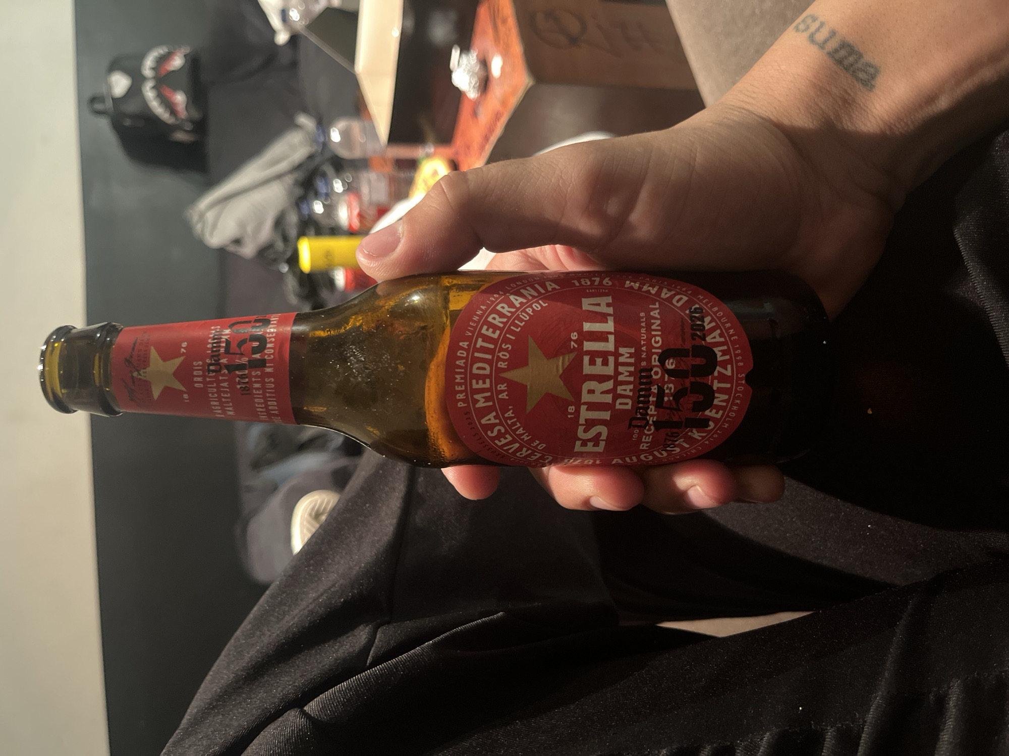 The image shows a person holding a bottle of Estrella Damm beer. The beer bottle label is clearly visible, displaying the brand name, location of manufacture, and other details such as "Cervesa Mediterrania", "1876" and "Recepta Original". The person's arm is visible, with a tattoo that reads "suma". The background is dark, with a partial view of what appears to be a table with various objects on it, including a backpack. The scene takes place indoors. The beer is linked to Barcelona, Spain.