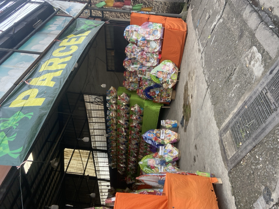 The image shows a storefront with a sign that says "PARCEL" in large yellow letters on a green banner with cartoon frogs. The storefront is located in Bojongsoang, Indonesia (latitude: -6.971, longitude: 107.637). The storefront is a semi-open space with a concrete floor. The storefront is selling gift baskets, which are arranged on shelves and in rows. The baskets are filled with various items, including food and other goods. Some of the baskets have price tags attached to them. There is an orange tarp covering the front of the store. There is a drain in the foreground. The image was taken on a clear day, with no people visible.