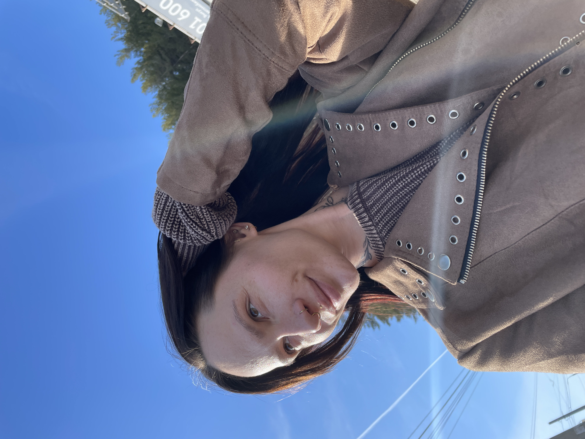 A woman poses for a selfie outdoors. She is wearing a brown jacket with silver eyelets and a patterned knit sweater. She has dark hair, a nose ring, and an ear piercing. Her hair is partially pulled back by a knit headband. The background shows a clear blue sky, some trees, and a sign. The location cannot be identified as Izhevsk, Russia from the information provided.