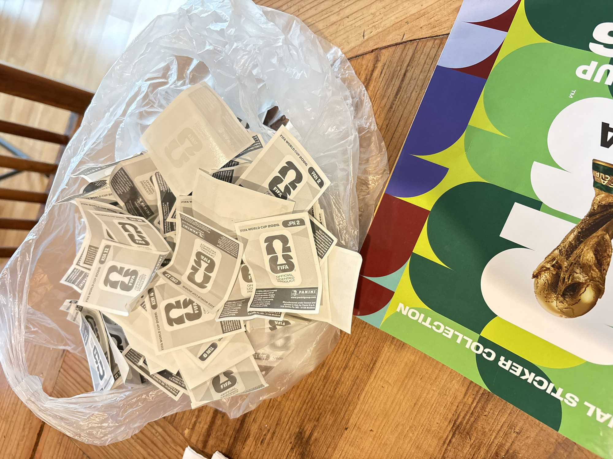 The image depicts a wooden table upon which are two main objects: a partially transparent plastic bag filled with numerous sticker backings from FIFA World Cup 2026 stickers, and a FIFA World Cup 2026 sticker collection album. The sticker backings in the bag are printed with the FIFA logo, the words "FIFA World Cup 2026," and various country codes. The album has a colorful graphic design featuring circles in shades of blue, green, yellow, and red. A partial image of the World Cup trophy is visible on the album cover. The background includes a portion of a wooden chair.