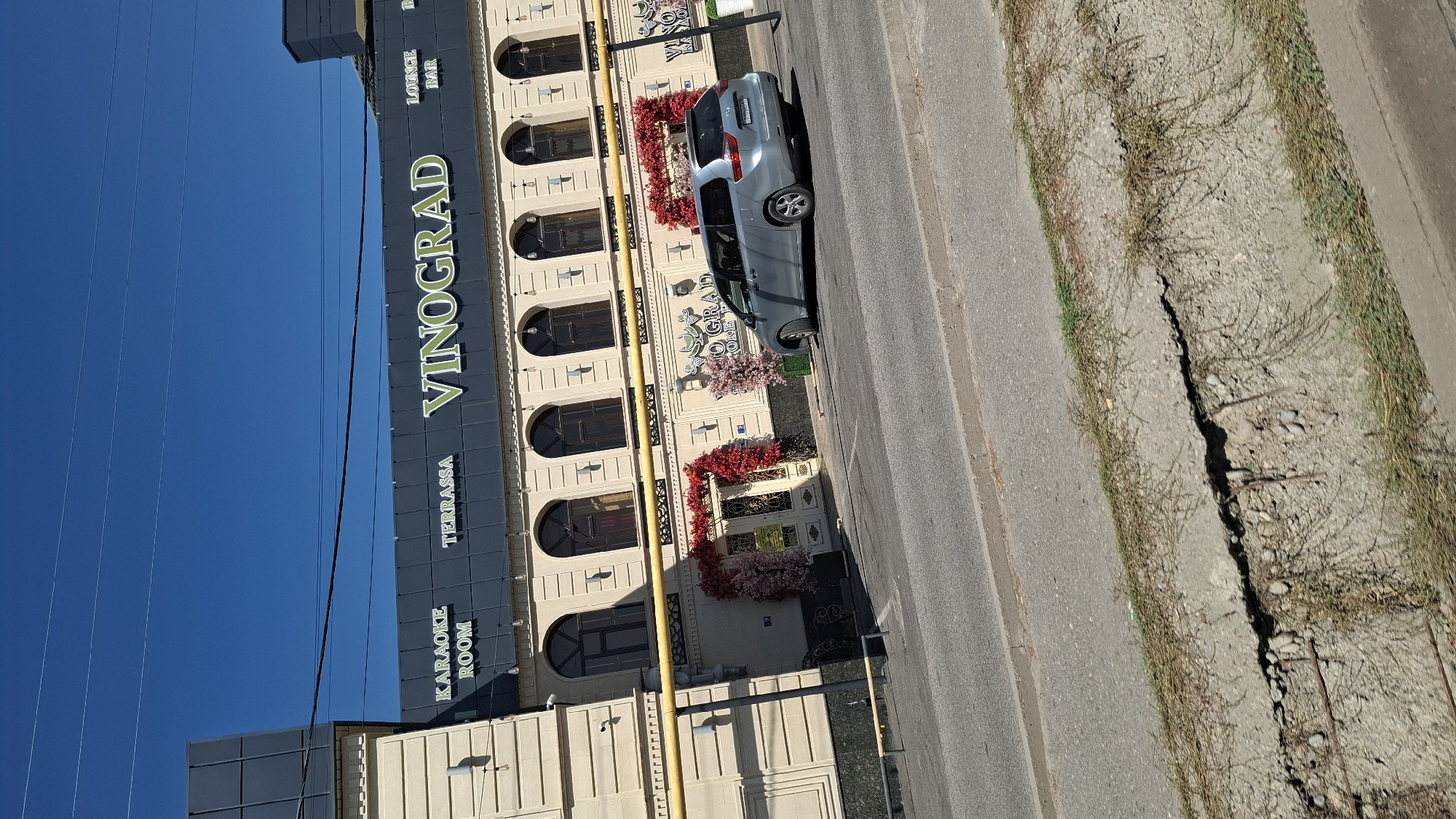 The image shows the exterior of a building with a sign that reads "VINOGRAD". The building appears to be a restaurant or entertainment venue, with "KARAOKE ROOM" and "TERRASSA" also visible on the sign. There are arched windows and decorative entrances on the building's facade. A silver minivan is parked in front of the building, and a street with a cracked concrete ditch runs along the foreground. The sky is clear and blue. The building is located in Shymkent, Kazakhstan.