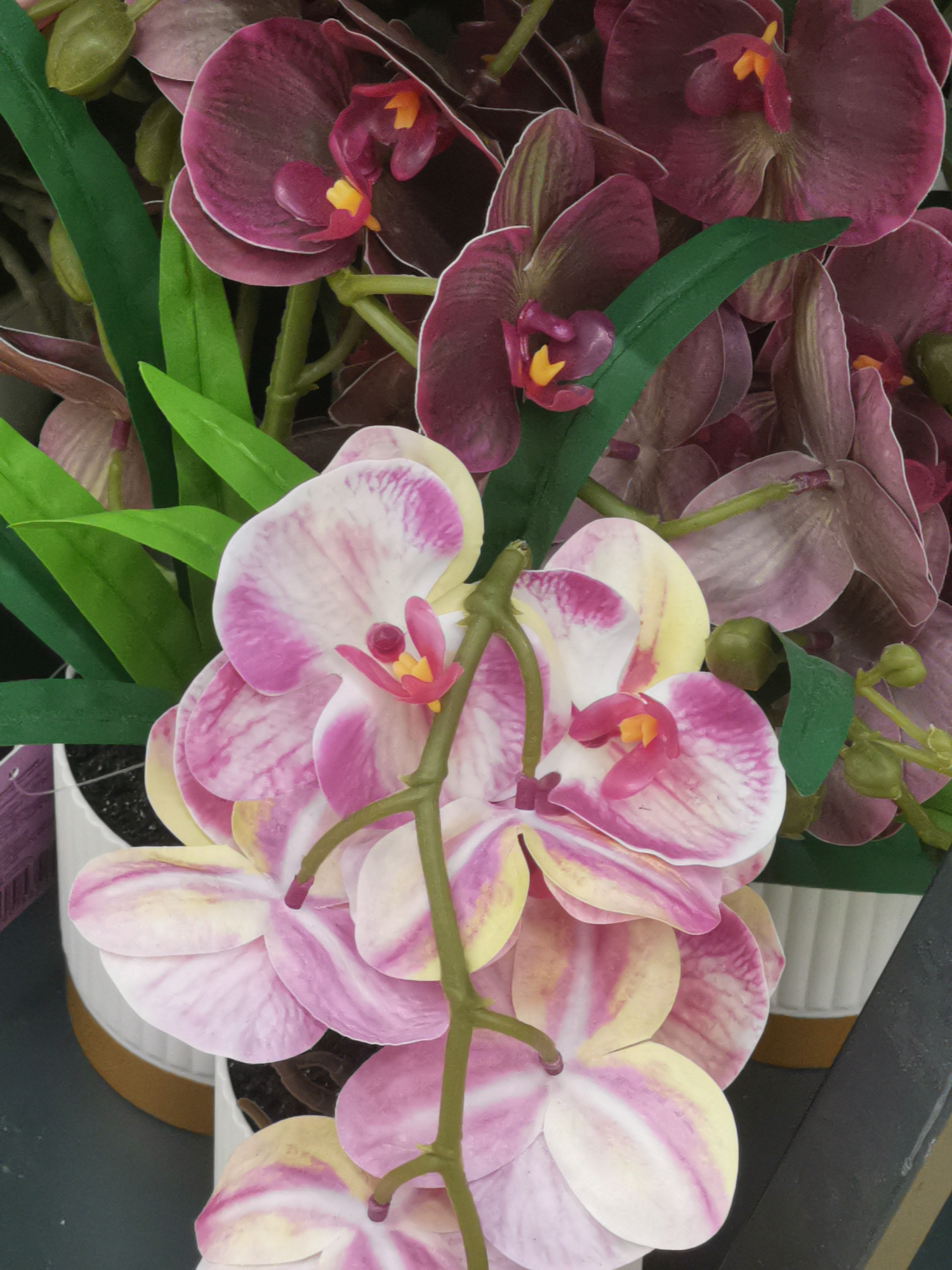 The image shows a close-up view of several artificial orchid flowers and leaves. The orchids are in varying shades of deep magenta and pale pink with yellow and red accents in their centers. Some of the flowers are fully open, while others appear to be buds. Green artificial leaves are interspersed among the flowers. The orchids are arranged in what appear to be white planters with a gold rim, and these are placed on a dark surface, possibly a shelf or table. The scene is brightly lit, suggesting an indoor setting such as a flower shop or home decor store. There are no people visible in the image. The image does not provide any information or context that would allow for its location to be identified as Cherkessk, Russia.