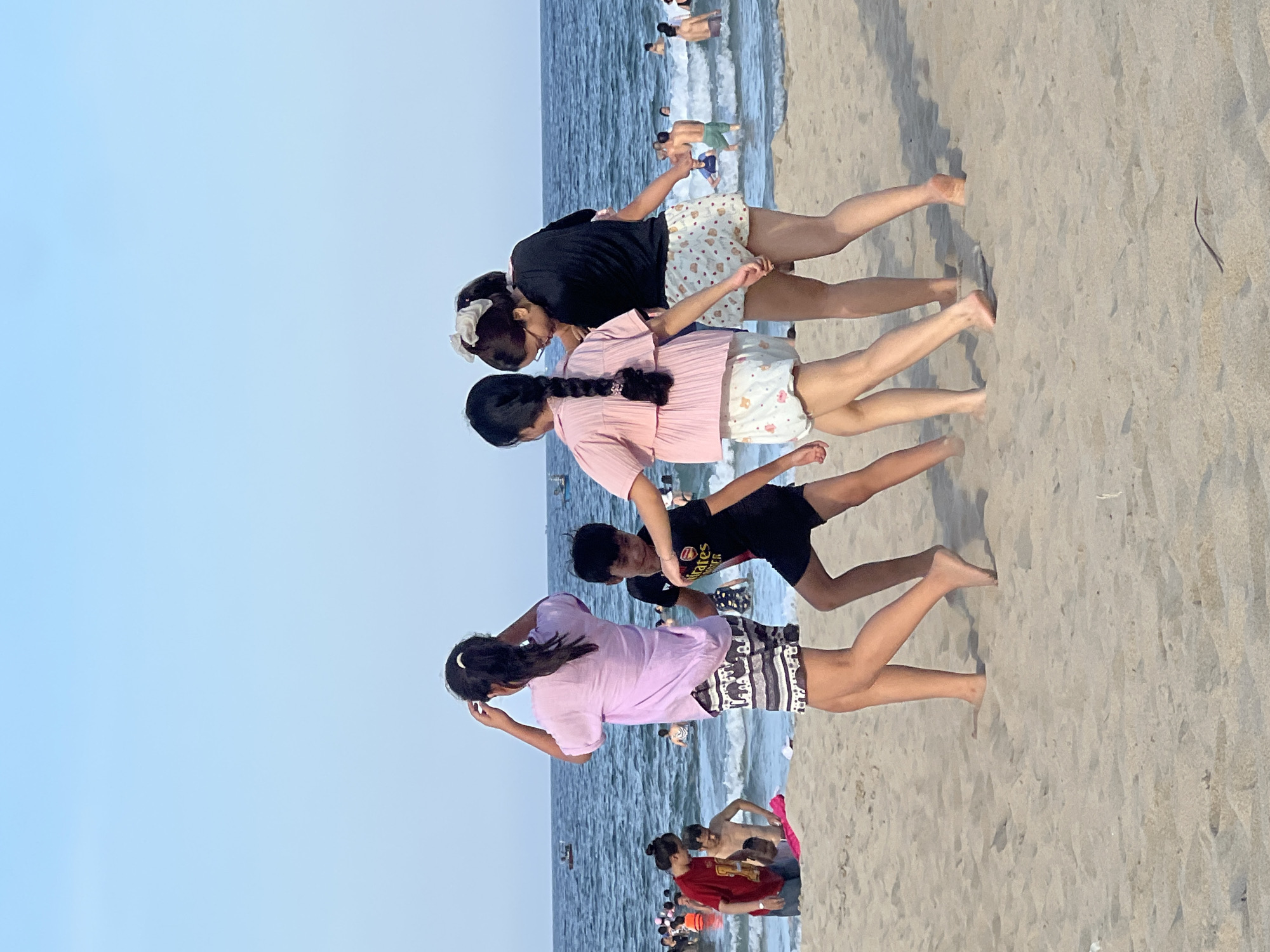 Four individuals, two young women and two children, are on a sandy beach with the ocean in the background. The two young women are on the right, one wearing a black shirt and patterned shorts, and the other wearing a pink top and similar patterned shorts. The younger girl has her hair in a braid and a white bow, while the other has her hair pulled back. They are holding hands, and one is carrying a very young child. To their left is a young boy wearing a black t-shirt with red lettering and black shorts. He is facing away from the camera. To the far left, another young woman in a light purple top and patterned shorts is adjusting her hair. The ocean is choppy with waves, and there are many people swimming and wading in the water. Several small boats are visible in the distance. The sky is clear and blue. The scene appears to be at My Khe Beach in Đà Nẵng, Vietnam, a popular tourist destination known for its sandy shores and clear waters.