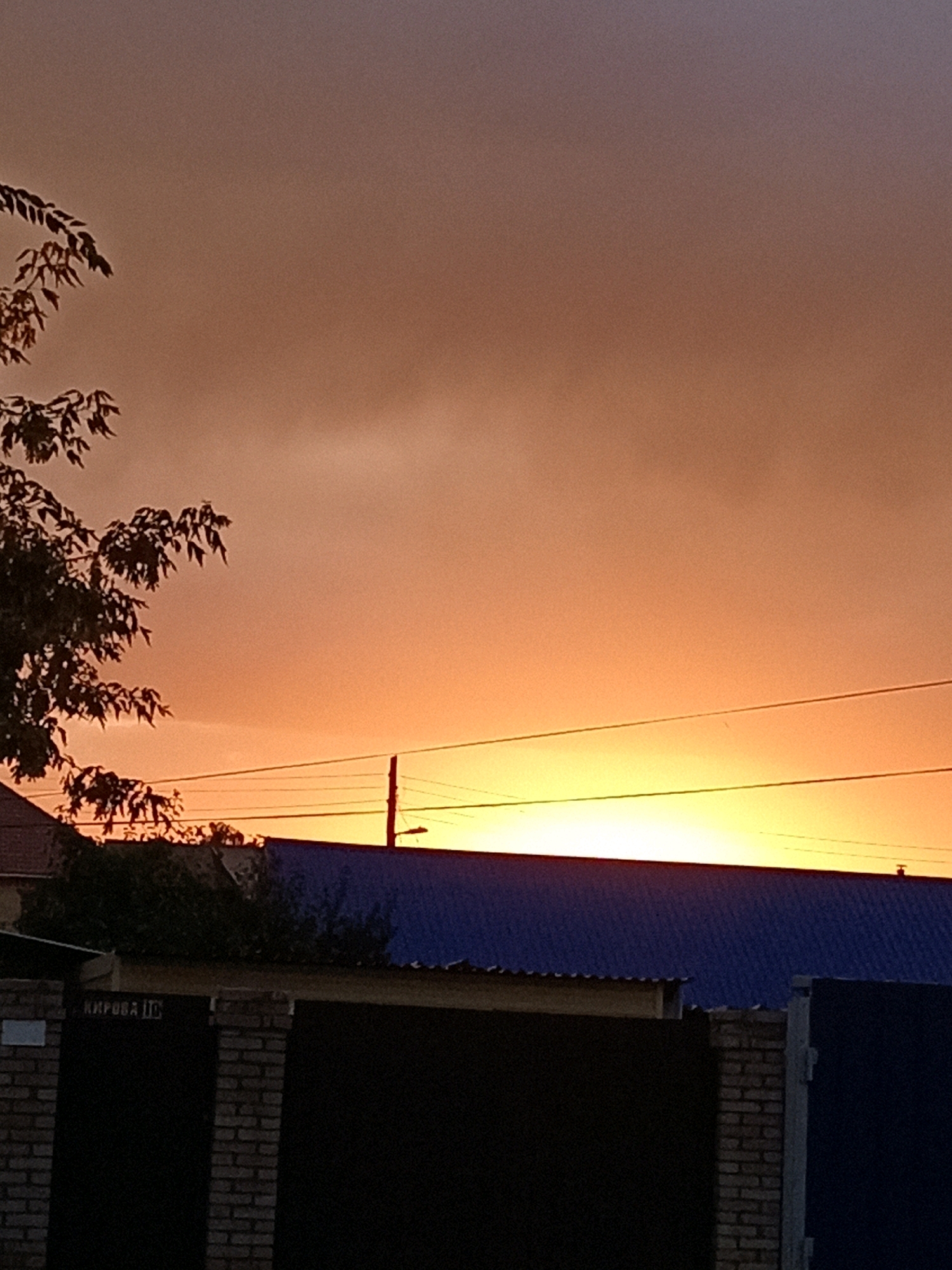 The image depicts a sunset over a residential area in Rudny, Kazakhstan. The sky is a gradient of warm orange and yellow hues, with darker, muted tones towards the top. Silhouetted against the bright sky are the branches of a leafy tree on the left, power lines stretching horizontally across the frame, and a utility pole. Below the sky, the roofs of several houses are visible. One prominent roof is covered in dark blue material. A black gate, constructed with brick pillars on the sides, occupies the foreground. The gate has a white plaque with Cyrillic text that appears to read "КИРОВА 10" (Kirova 10), likely indicating a street address. The overall scene is one of a quiet evening in a suburban or semi-urban environment.