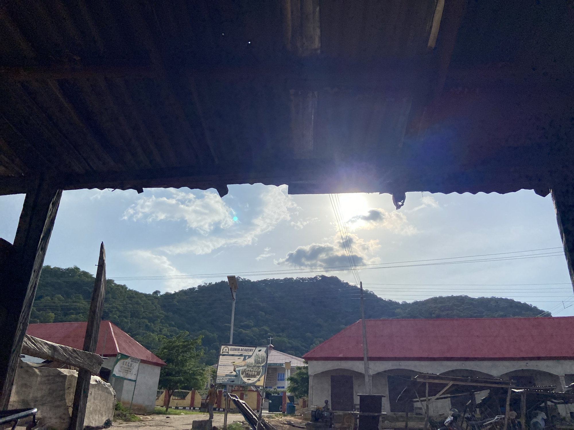 The image captures a view of a sunlit sky with clouds, peeking through the corrugated iron roof of a structure in the foreground. In the middle ground, two buildings with red roofs are visible. One of these buildings has a sign that reads "ELOHIM ACADEMY." A forested hill forms the backdrop. The scene appears to be in the city of Mararaba, Nigeria, suggested by the presence of the Elohim Academy. The overall impression is one of a bright, possibly warm day in a semi-urban or rural setting.