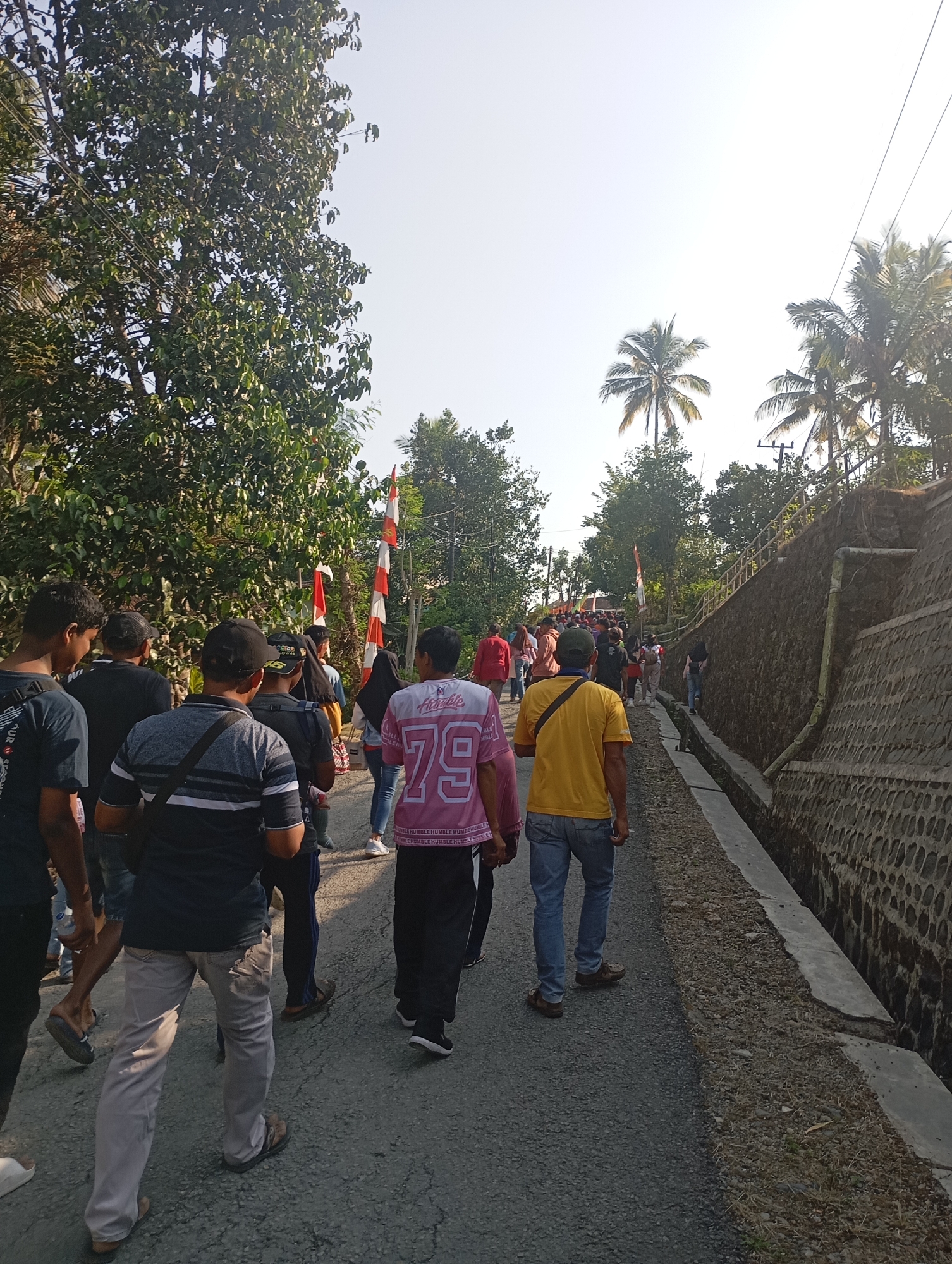In the city of Ketepung, Indonesia, a group of people walks along a paved road bordered by a stone wall on the right and trees on the left. The sky is bright and clear. Several individuals are visible in the foreground, walking away from the camera. One person in a pink shirt with the number 79 is prominent. Another person in a yellow shirt is also in the foreground. The road appears to be winding uphill. Red and white Indonesian flags are displayed on poles along the left side of the road, suggesting a celebration or event. Palm trees are visible in the background, indicating a tropical climate. The overall scene depicts a procession or gathering on a sunny day in a semi-rural Indonesian setting.