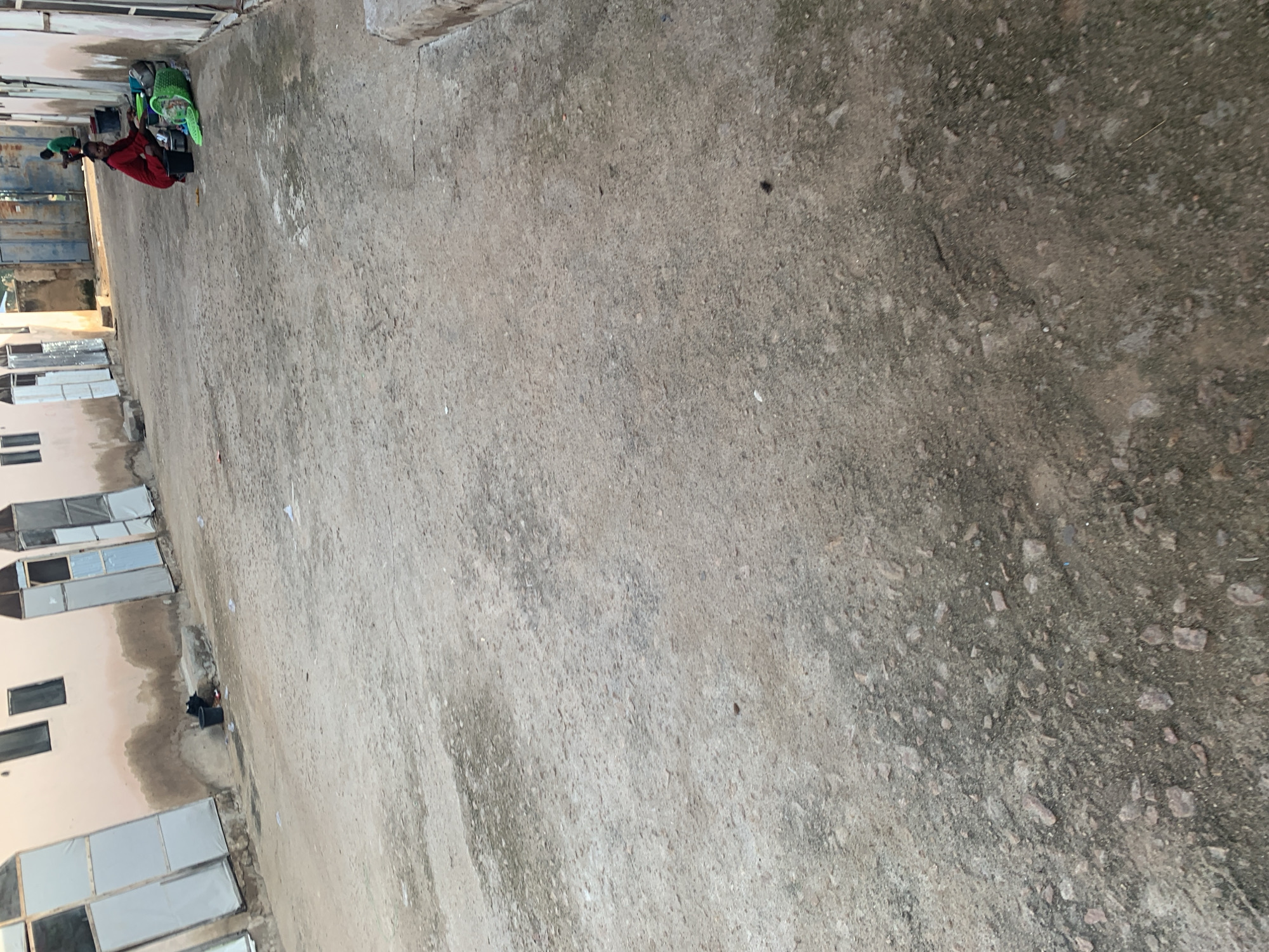 In Jauro Yinu, Nigeria, a street scene unfolds with a building featuring light pink walls and multiple white doors or entrances. To the right, a young woman in a red dress is crouching, engaged with items on the ground, including a green basket and various utensils. Nearby, a child in a green shirt is also on the ground. The ground surface is a mixture of packed dirt and gravel. The overall setting appears to be an outdoor residential or communal area.