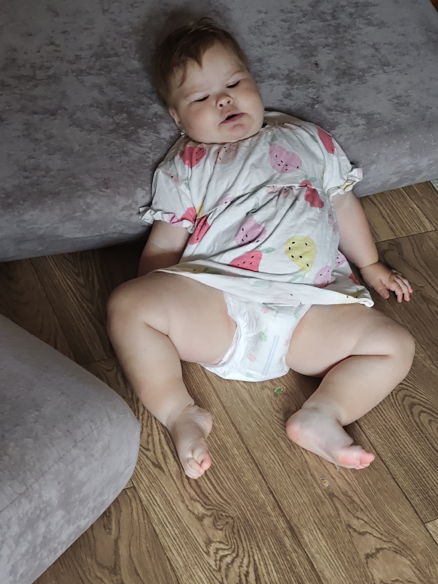 A baby is lying on the floor, next to a grey couch. The baby is wearing a white dress with a strawberry print and a diaper. The floor is made of wooden planks. The baby appears to be sleeping or resting, with eyes closed and mouth slightly open. The lighting suggests it is daytime, and the indoor setting implies a home environment. The image is taken from a high angle, looking down at the baby. There is no visible text in the image. The setting is likely a living room or bedroom in Chaikovsky, Russia.