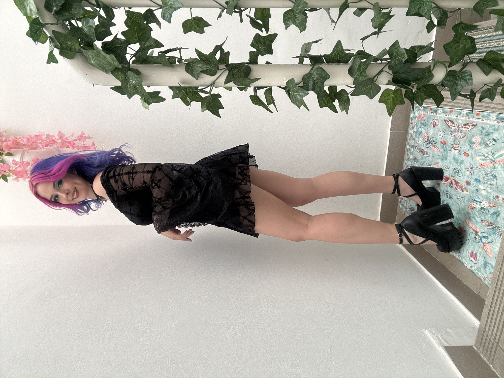 A young woman with vibrant, dual-toned purple and pink hair is captured in a full-body shot, appearing to lie on her back and gaze upwards towards the camera. She is smiling broadly, conveying a cheerful and confident expression. She wears a sheer black lace mini-dress or babydoll with long sleeves, and black platform high heels.
The setting appears to be indoors, against a plain white wall or floor that acts as a minimalist background. Above her head (in the correctly oriented view), a white bar or pipe is adorned with artificial green ivy vines. Nearby, a cluster of pink artificial flowers, possibly cherry blossoms, adds a decorative touch. To her right, a colorful patterned rug or mat featuring butterflies, flowers, and other motifs in shades of blue, green, pink, and orange is visible on the floor, next to what appears to be a raised step or edge.
The lighting is bright and even, suggesting an indoor daytime setting. The woman is posing, with her body elongated, emphasizing her legs and attire. There is no visible text in the image.