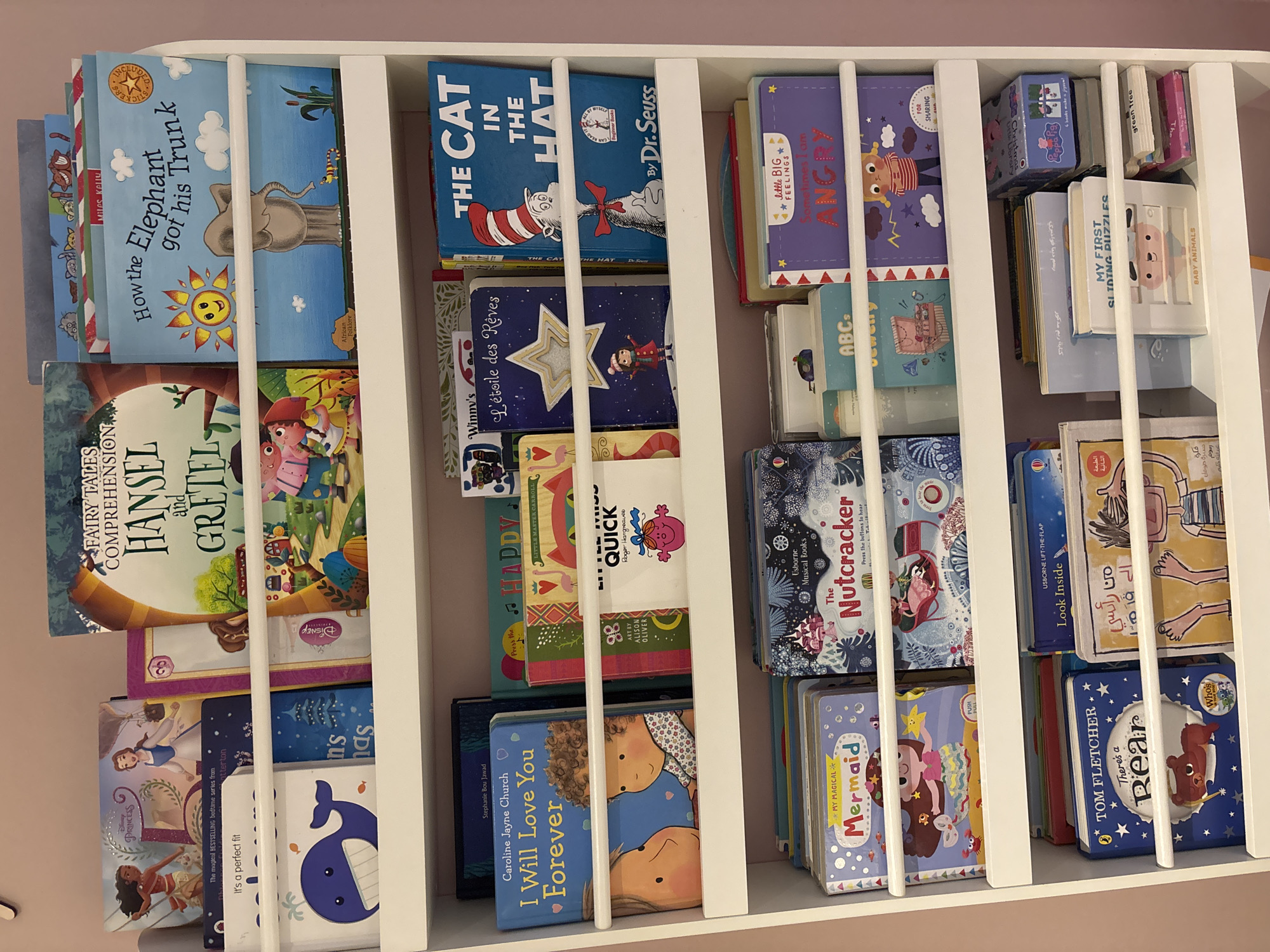 The image shows a white, three-tiered bookshelf filled with children's books. The bookshelf is positioned against a pale pink wall, suggesting an indoor setting, likely a child's bedroom or playroom. The books are varied in size and cover, with many featuring bright illustrations and titles in English. Some notable titles visible include "How the Elephant got his Trunk," "The Cat in the Hat," "The Nutcracker," and "My Magical Mermaid." The books are arranged in a neat, organized manner, with some standing upright and others lying flat. There are no people or animals visible in the image, and no specific actions are taking place. The environment appears clean and well-lit. The image does not provide any information that would suggest a location in Dubai, United Arab Emirates; it is a generic indoor scene.