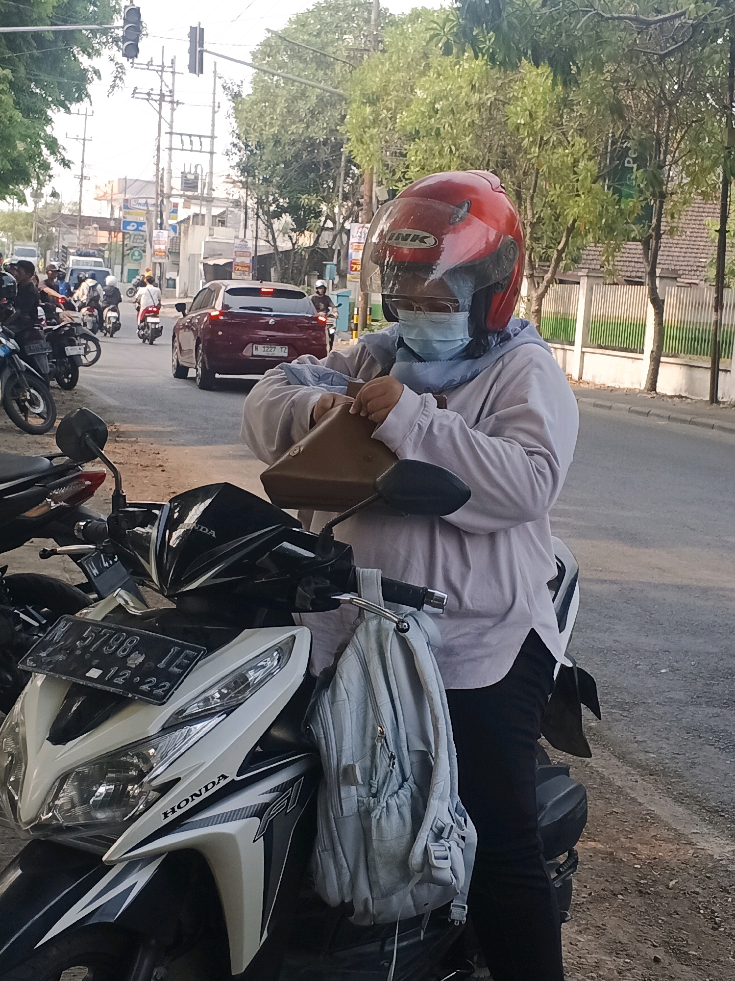 A person wearing a red helmet and a light-colored long-sleeved shirt with a blue headscarf and a surgical mask is seated on a white Honda motorcycle. They are holding a brown bag in their hands, appearing to be placing it into a compartment or bag attached to the motorcycle. A light blue backpack is also attached to the back of the motorcycle. The motorcycle is parked on a street in Krian, Indonesia, with other motorcycles and a red car visible in the background. Traffic lights and utility poles are also present, indicating an urban environment. The time of day appears to be late afternoon or early evening due to the lighting conditions.