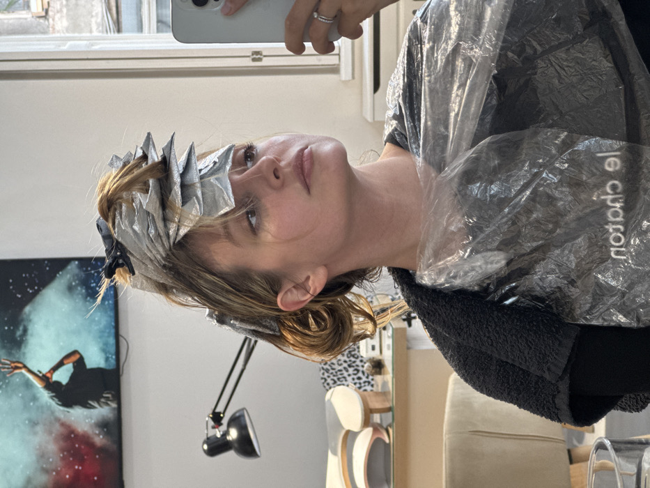 A female individual, appearing to be in her 20s or 30s, is depicted in a salon environment located in Tbilisi, Georgia. Her light brown hair is actively undergoing a chemical treatment, with numerous sections individually wrapped in silver foils and secured with dark clips, indicating a highlighting or coloring process. She is covered by a black, textured towel draped over her shoulders and a clear, disposable plastic cape with the inverted text "noitpo le" (Le Option) printed on it, protecting her clothing. Her right hand holds a light-colored smartphone, positioned to capture a selfie, and a silver ring is visible on her ring finger. Her gaze is directed slightly upwards and to the right. The background on the left features a white wall with a mounted flat-screen television displaying an image of a person with raised arms amidst red and blue smoke. Below the television, a black adjustable desk lamp is visible. Further left, a light-colored upholstered chair and a counter with decorative objects, including a distinctively curved wooden piece and a hint of leopard print, are present. To the right, a window provides a view of an older building's exterior, characteristic of an urban setting, alongside a white cabinet. The specific location is identified as "Le Option Salon" in Tbilisi, Georgia, based on the text on the protective cape.
