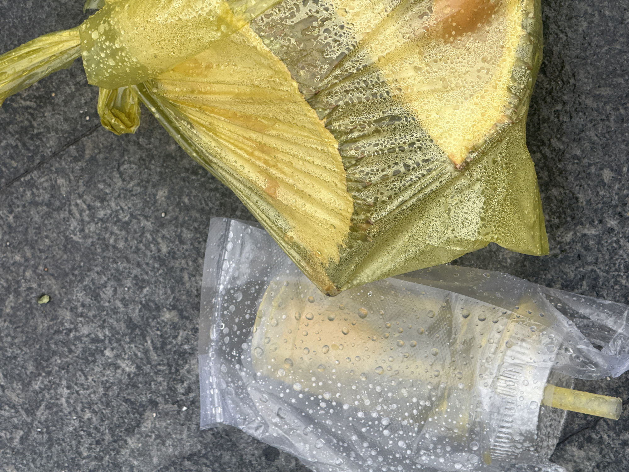 The image displays two plastic bags on a grey, textured surface, likely asphalt. One bag is clear and contains a cylindrical object with a ridged silver base and a yellow stick protruding from it. The second bag is bright yellow and appears to hold several pieces of a pale yellow food item. Both bags are covered in condensation. The setting is identified as Phu Thuy Ward, Vietnam. There are no people visible in the image.