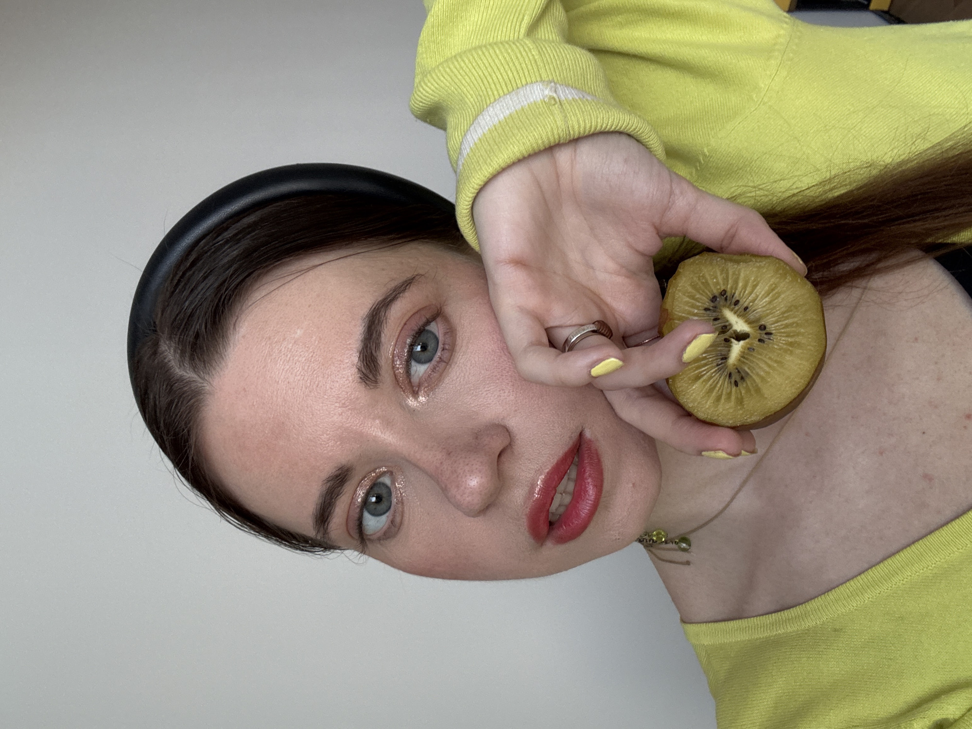 A woman is holding a slice of golden kiwi near her face. She has light skin, light blue eyes, and dark hair. She is wearing a black headband, a yellow sweater with white stripes on the cuff, and yellow nail polish. She also has a silver ring on one of her fingers and a delicate gold necklace. The background is a plain white wall. There is no indication of the city of Moscow, Russia.