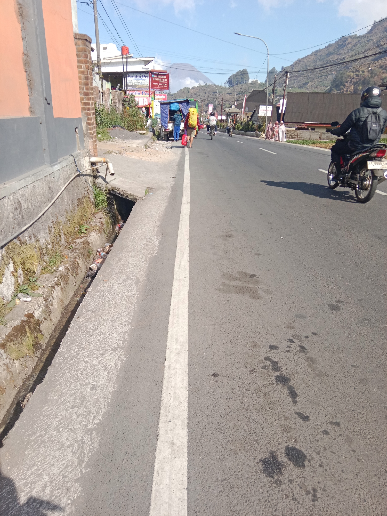 The image depicts a street scene in Parikesit, Indonesia, under a clear sky with scattered clouds. The road, paved with asphalt, has a white line marking the lane separation. To the left of the road is a concrete drainage ditch containing scattered refuse. Adjacent to the ditch, a building with an orange and gray facade is visible, with a brick portion and a downspout. Further down the street, several individuals are present. Two people, one with a yellow backpack and the other with a blue jacket, walk along the road's edge. Behind them, a small truck with a blue tarp covers its load is parked. A motorcyclist wearing a black jacket and helmet rides a motorcycle with license plate "AA 6928 LM" on the road. In the background, several buildings line the street, and a mountainous landscape with vegetation can be seen, with a smoking volcano or large industrial chimney in the distance. The overall scene conveys a sense of everyday life in a developing urban area, with elements of infrastructure, commerce, and natural surroundings.