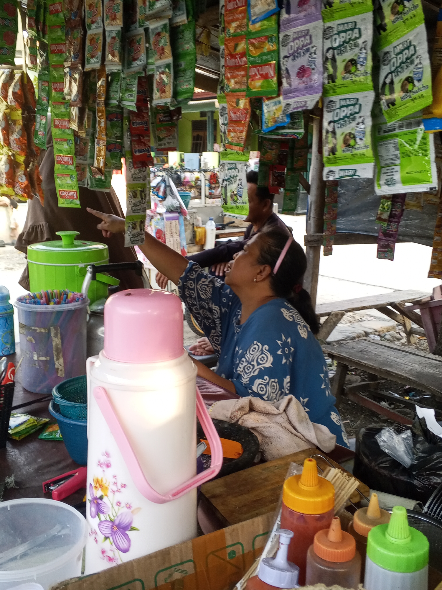 The image shows a street vendor stall in Ciruas, Indonesia, with numerous small packets of food or drink products hanging from the ceiling. In the foreground, a large white and pink thermos with a floral design sits on a table, next to a clear plastic container filled with colorful straws. Several bottles of condiments, including what appear to be ketchup and chili sauce, are also visible on the table. Behind the thermos, a blue plastic bucket and a green plastic container are present. Two individuals are partially visible; a woman in a blue patterned top is seated and looking upwards, and another person in a brown headscarf is standing and pointing towards the hanging products. The stall appears to be outdoors, with wooden structures and benches suggesting a simple, open-air setup. The overall scene depicts a busy, everyday transaction at a local market or roadside stall.