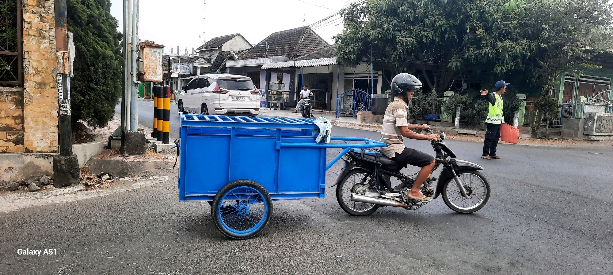 In Tangkil, Indonesia, a man wearing a brown and white striped shirt, black shorts, and a dark helmet rides a black motorcycle. The motorcycle is towing a large, bright blue trailer with two wheels. In the background, a white SUV is parked on the side of the road, and several buildings with tiled roofs line the street. A second man, wearing a bright yellow safety vest, dark pants, and a blue cap, stands on the side of the road holding an orange bag. He is gesturing with his left hand. The environment appears to be a semi-urban street with trees and sidewalks. The image captures a moment of daily life and traffic on a street in Tangkil.