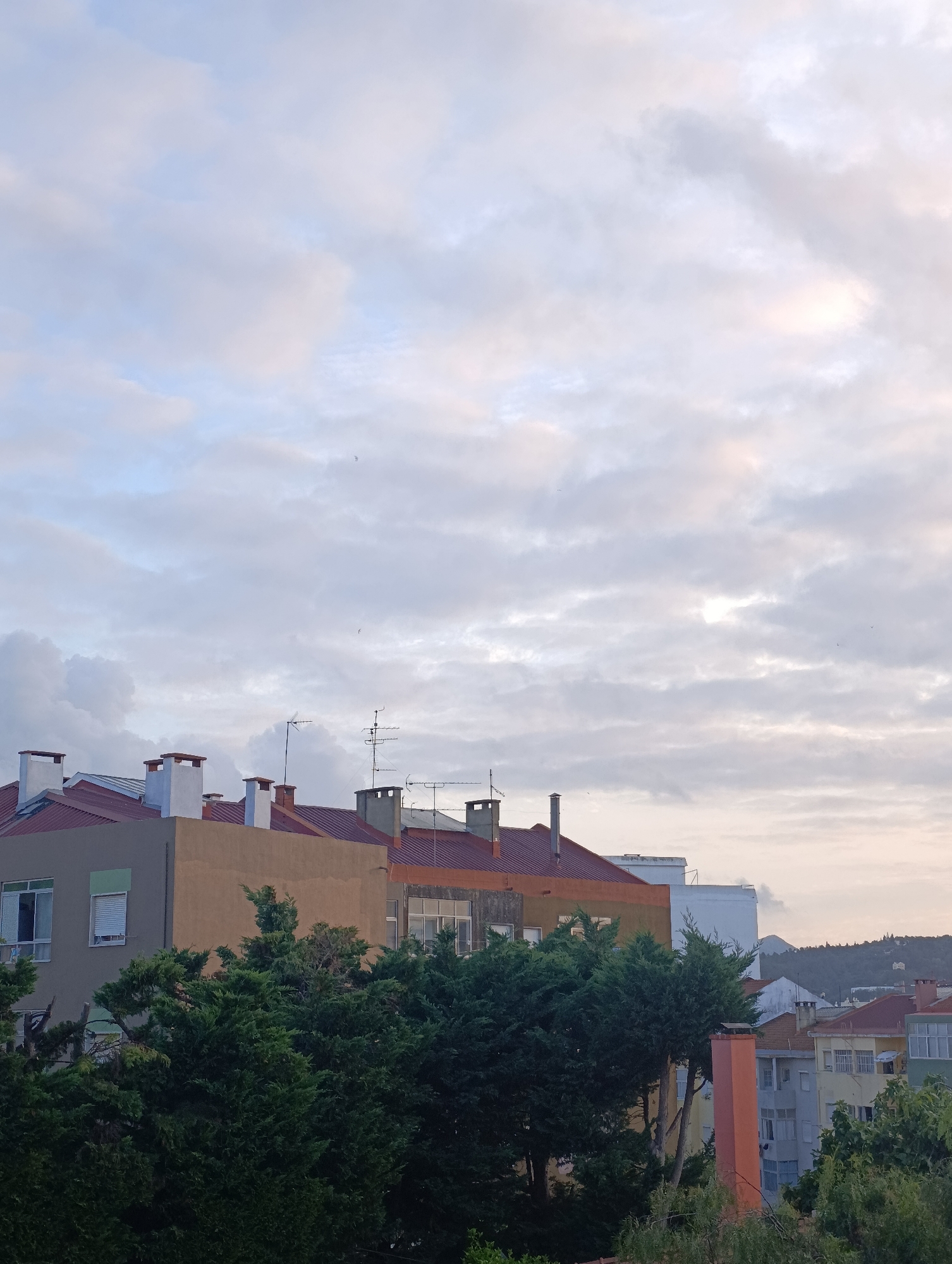 The image depicts a view of buildings and trees in Setúbal, Portugal, under a cloudy sky. The buildings have red-tiled roofs and are of varying heights and colors, including tan, orange, and light gray. Chimneys are visible on several rooftops. Dense green trees occupy the foreground and midground, partially obscuring some of the lower levels of the buildings. In the background, a distant hillside with more buildings and vegetation is visible. The sky is filled with soft, diffused clouds, suggesting either early morning or late afternoon lighting conditions. There are no people or animals visible in the image.