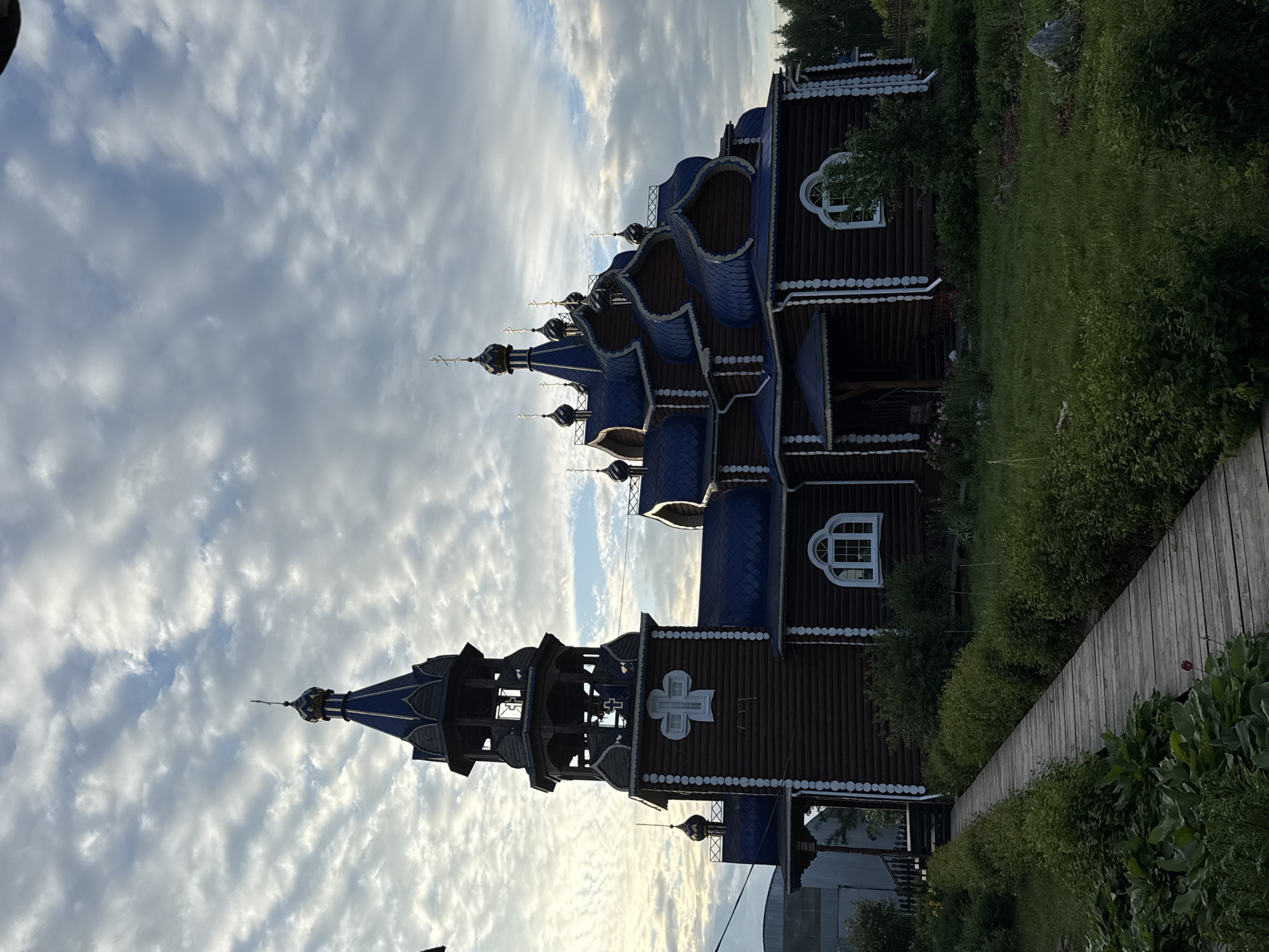 The image depicts an exterior view of a traditional Russian wooden church with blue onion domes, located in the village of Kukuy, Russia. The church is constructed primarily from dark wood logs and features ornate white trim around the windows and edges. A tall bell tower with multiple levels is attached to the left side of the main structure. Several smaller domes are visible on the roofline, each topped with a cross.
A wooden walkway, made of planks, leads from the foreground towards the entrance of the church, bordered by lush green vegetation and bushes. The ground is covered with grass, and in the background, trees are visible. The sky above is filled with dramatic clouds, suggesting it might be early morning or late afternoon. There are no people or animals visible in the scene. The overall impression is one of a serene, rural religious setting.