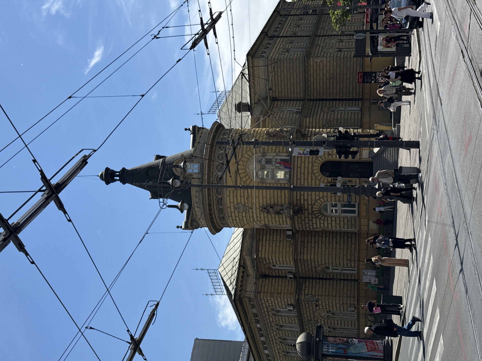 The image shows a street scene in Vracar Urban Municipality, Serbia. A tall, ornate building with a distinctive tower dominates the view. A Serbian flag is visible on the tower. Pedestrians are crossing the street at a zebra crossing, with a mix of clothing styles and ages. Overhead tram lines crisscross the sky. Street lighting and traffic lights are also present. The sky is blue with some clouds.