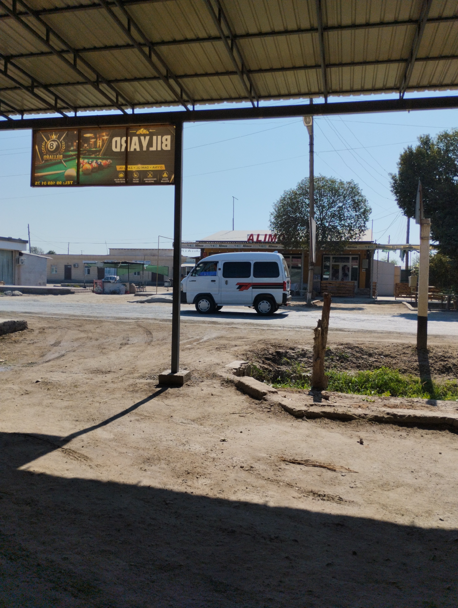 The image depicts a sunlit scene in Yangibazar, Uzbekistan, likely a street or an outdoor establishment. In the foreground, a patch of dry, dusty ground is visible, with tire tracks imprinted in it. A large shadow of an overhead structure falls across the lower portion of the image.
In the middle ground, a white van with red and black stripes is parked on a paved surface. To the left of the van, a sign advertising "BILYARD" (Billiard) is prominently displayed, featuring an image of billiard balls and cues. The sign appears to be in a foreign language, possibly Uzbek or Russian, given the location. Beyond the van, a building with an awning and outdoor seating suggests a commercial establishment, possibly a cafe or restaurant, with the name "ALIMO" visible on its facade. A street lamp stands to the right of the van.
The background reveals a clear blue sky and trees, indicating an outdoor setting in a town or city. The overall impression is one of a warm, daytime scene in a developing urban environment. No individuals are clearly visible in the image.