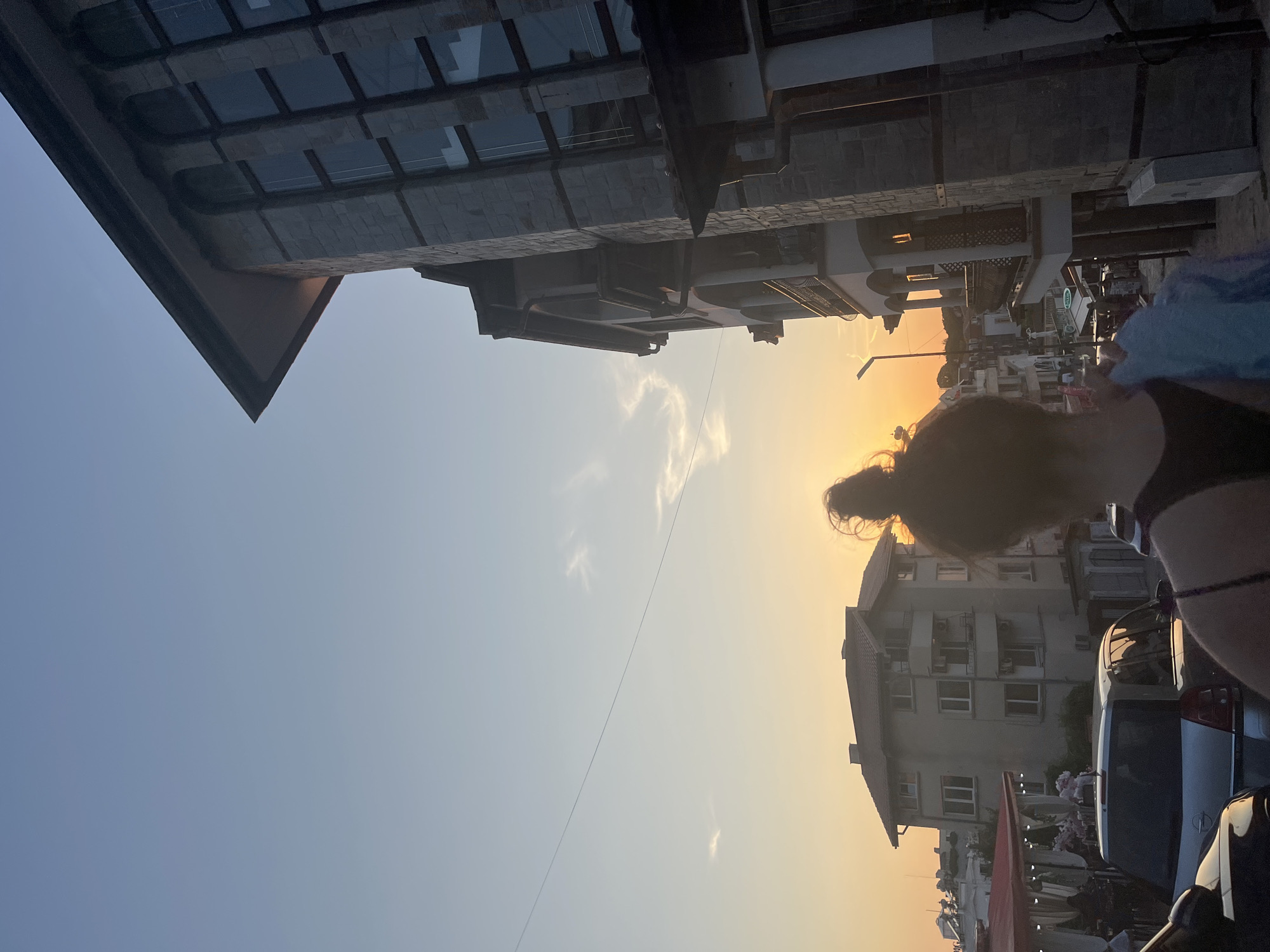 The image captures a street scene in Lozenets, Bulgaria, during what appears to be sunset. The sky is a gradient of light blue to a warm orange hue, with a few wispy clouds visible. A prominent building with stone facade and large windows dominates the right side of the frame, its architecture suggesting it may be a hotel or apartment complex. To the left, a multi-story residential building with a tiled roof is visible, alongside what looks like an outdoor dining area with red awnings. Several cars are parked on the street, including a silver hatchback in the foreground. In the center of the image, a person with their back to the camera is seen, their hair tied up in a bun. They are holding a light blue item, possibly a piece of clothing. The overall impression is a relatively quiet street in a town by the sea, bathed in the soft light of the setting sun.