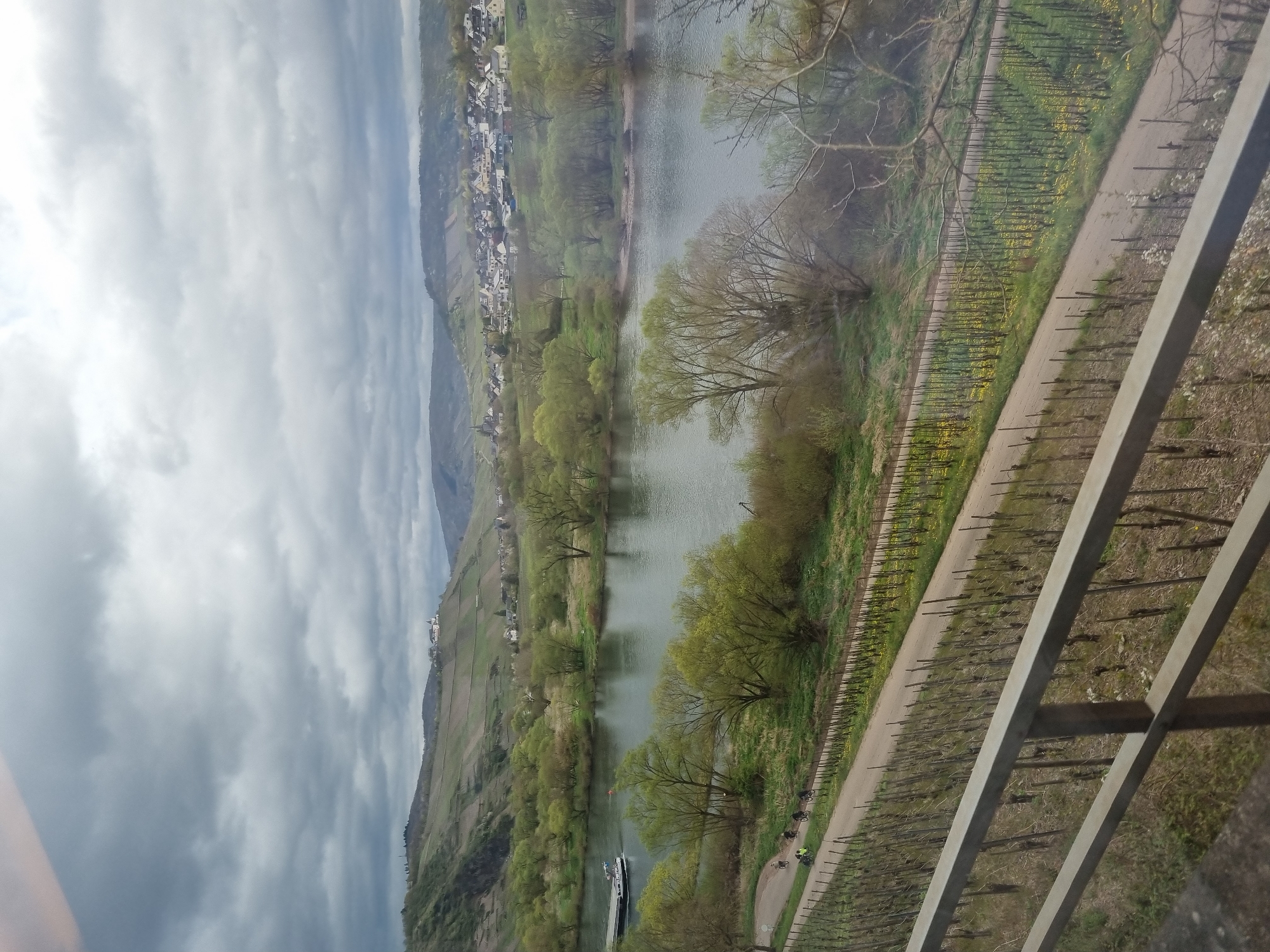 The image shows a view overlooking the Moselle River in Cochem, Germany. A small barge is traveling along the river. The steep slopes on either side of the river are covered with vineyards. A road runs alongside the river on one side, with several cyclists visible. The sky is overcast with dark clouds. A metal railing is visible in the foreground, partially obscuring the view.