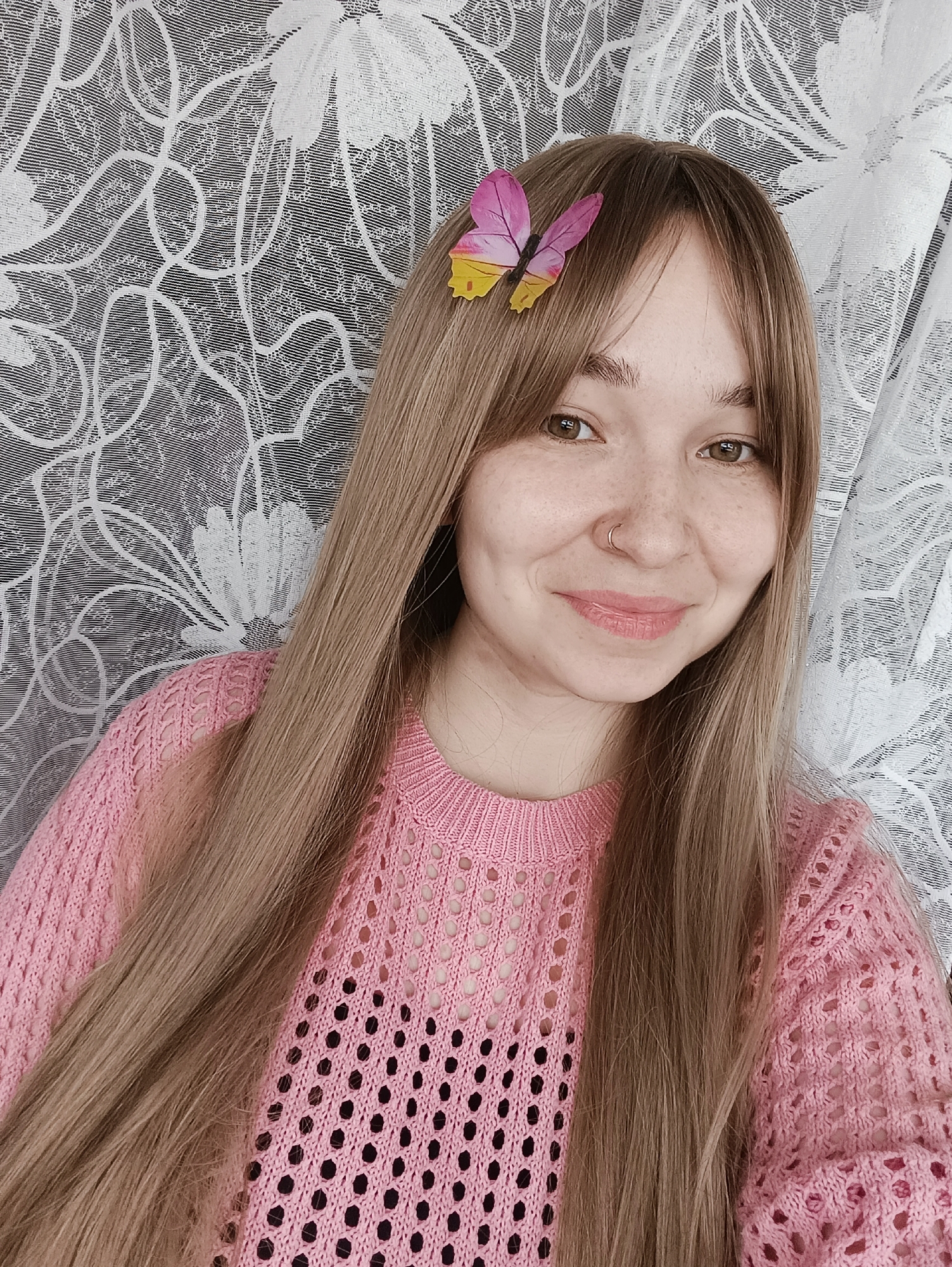 A young woman poses for a selfie. She has long, light brown hair with bangs, brown eyes, and a nose ring. She is wearing a pink sweater with a crochet-like pattern. A butterfly hair clip adorns her hair. The background features a patterned wall covering or curtain. The location is not discernible from the image.