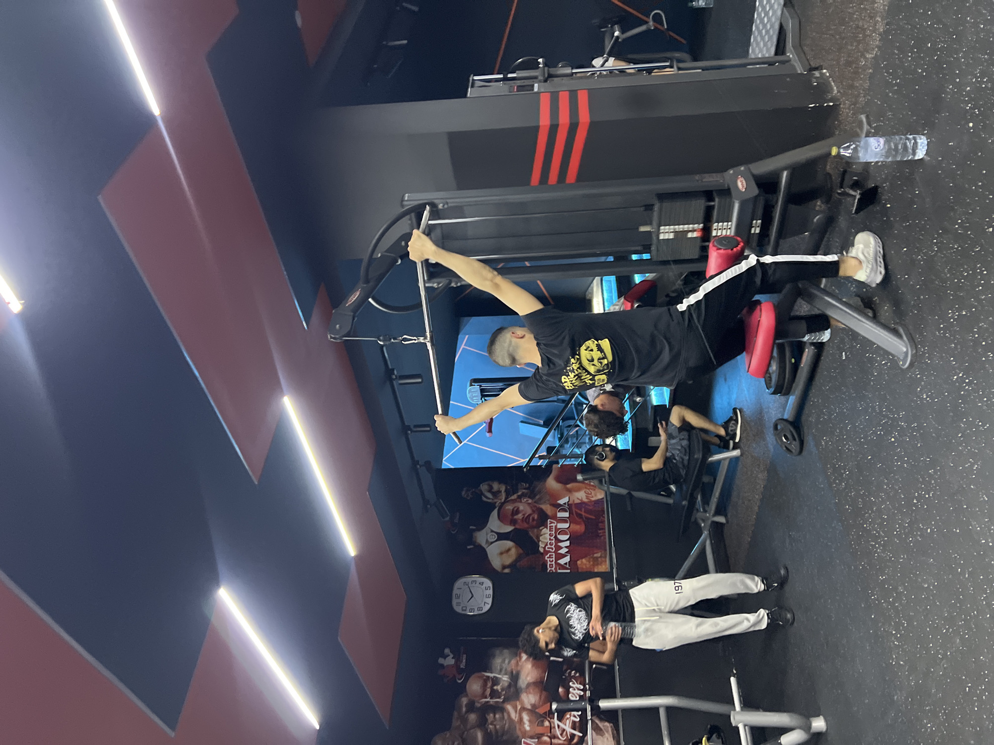 The image captures an active scene inside a modern gym or fitness center located in M'Diq, Morocco. The gym features a dark aesthetic with walls partially adorned with geometric patterns in black and a reddish-brown hue, illuminated by linear LED strip lights. The floor is covered with a speckled dark gray rubber mat.
Several individuals are present, engaged in various fitness activities. In the central area, a man wearing a black t-shirt with a yellow graphic and dark pants with white stripes is actively using a cable machine, extending one arm upwards. A clear plastic water bottle is placed on the equipment frame near his feet. Nearby, another person in a black t-shirt and light gray sweatpants, which have the number "197" printed on them, stands casually. Further back, another individual is seated on a weight bench.
The gym is equipped with standard workout machinery, including cable stations and weight benches. Decorative elements include large wall posters featuring images of bodybuilders or athletes, with visible text such as "Dach Jeremy" and "TAMOUDA". A white circular clock on the wall indicates the time is approximately 11:10. Other partial text elements like "Fu ess" and "Tones" are also visible, likely part of gym branding or other posters. The overall atmosphere is typical of a fitness facility, with people focused on their workouts.