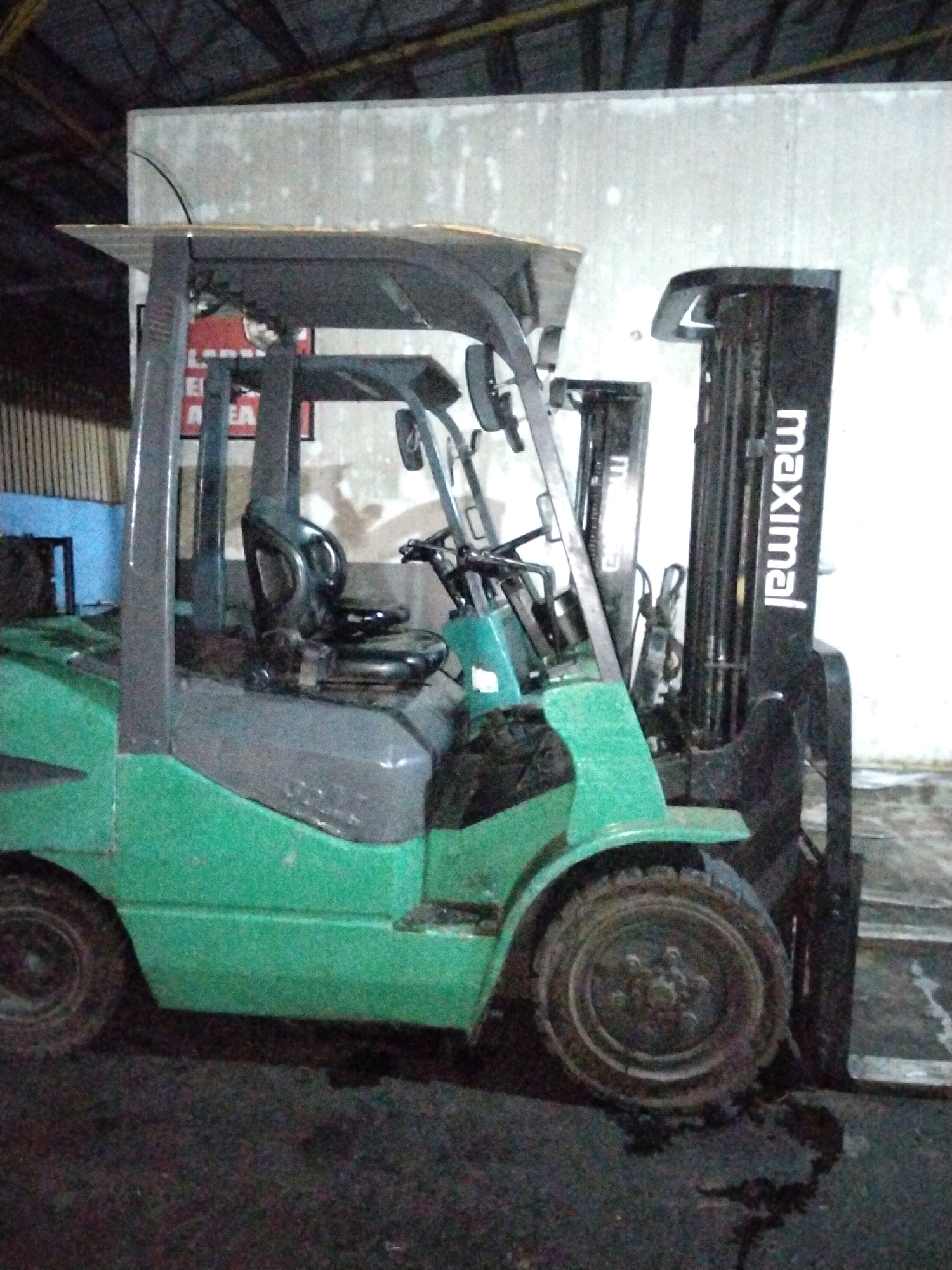 A green and black forklift is parked in what appears to be a warehouse or industrial area. The forklift has a black overhead guard and a black seat. The brand name "Maximal" is visible in white lettering on the black mast of the forklift. The forklift is in the foreground and takes up a significant portion of the image. The background is dimly lit and shows a concrete wall and possibly some industrial shelving. The ground is dark and appears to be concrete or asphalt, with some dark liquid spilled on it in front of the forklift. The overall lighting is dim, suggesting it may be nighttime or indoors with artificial lighting. There are no people or other vehicles visible in the image. The context of Moro, Indonesia, is not evident from the image itself.
