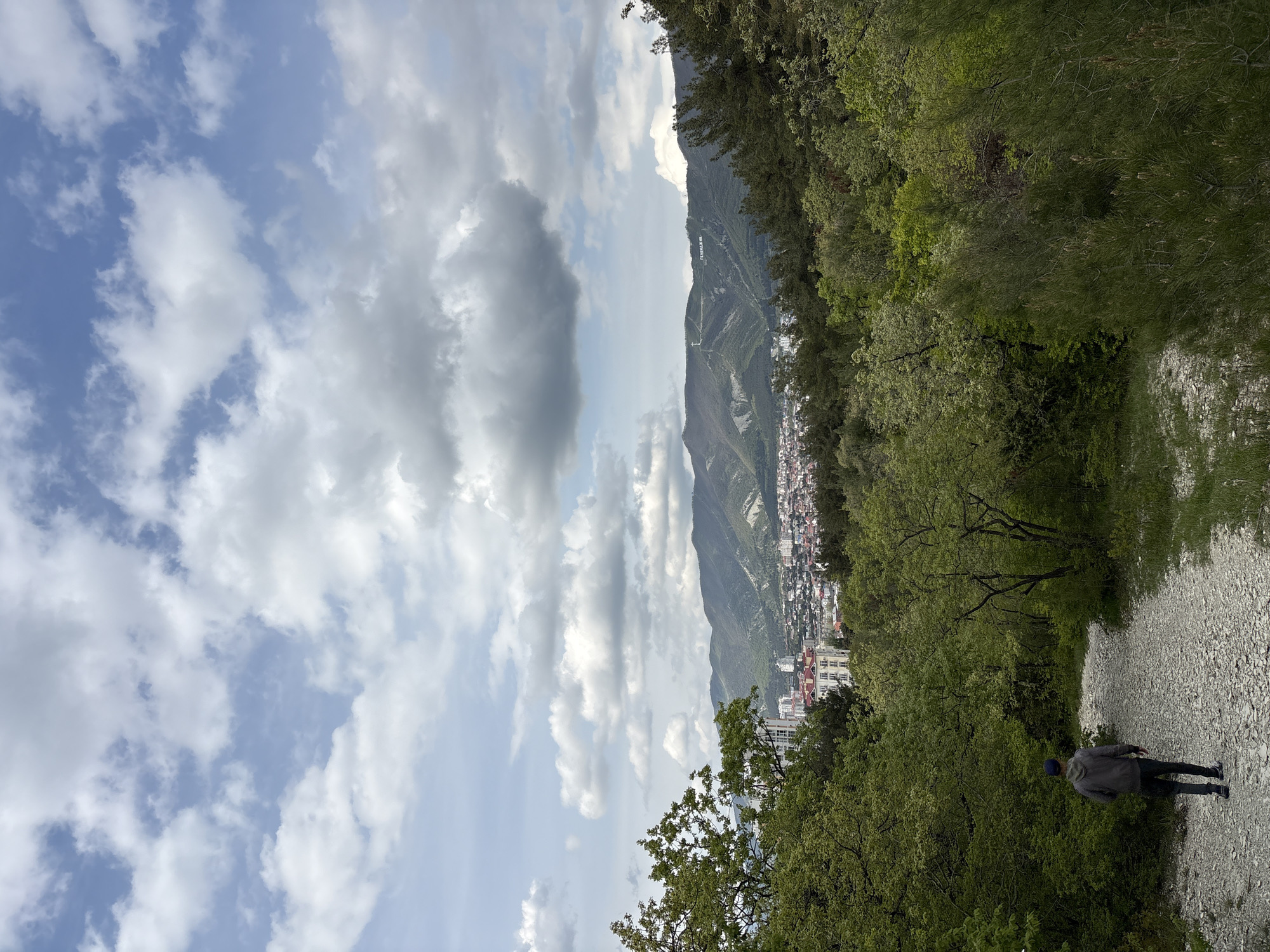 The image depicts a panoramic view of a mountainous landscape overlooking a town, taken from a slightly elevated vantage point. The perspective is from a gravel path, where a lone individual, dressed in a grey jacket, dark pants, and a blue cap, is walking away from the viewer towards the middle ground. Lush green trees and dense foliage frame the path on both sides, creating a natural border.
In the mid-ground and background, a sprawling town or city with numerous buildings, many featuring red rooftops, stretches along the base of impressive, verdant mountains. The mountains dominate the horizon, with some peaks appearing rugged and others covered in trees. On one of the more distant mountain slopes, prominent white letters spell out "ГЕЛЕНДЖИК" (Gelendzhik) in Cyrillic, serving as a clear location identifier.
The sky is a bright blue with scattered, fluffy white and grey clouds, suggesting a partly cloudy day. The illumination indicates it is daytime, likely late morning or early afternoon. The overall scene conveys a sense of natural beauty combined with urban development, characteristic of a resort town nestled by mountains and presumably a coast, though the body of water is not directly visible.