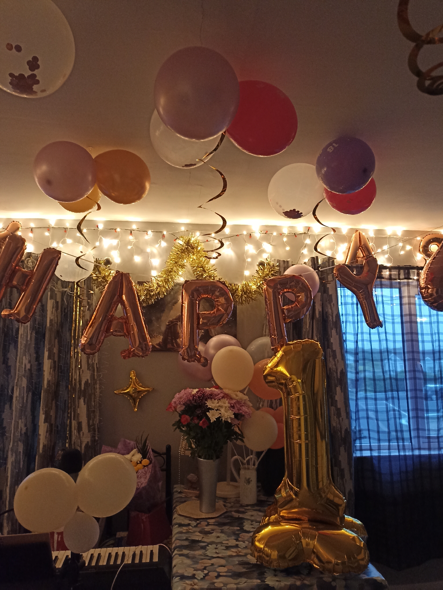 The image depicts a room decorated for a celebration, likely a birthday, given the prominent "HAPPY 1" balloon formation. Numerous balloons in various colors, including white, pink, red, orange, and purple, are scattered across the ceiling and suspended by ribbons. Some balloons are clear with confetti inside. A string of fairy lights is draped across the ceiling, adding to the festive atmosphere.
The word "HAPPY" is spelled out in large, rose-gold foil balloons. Following this, a large, gold foil balloon shaped like the number "1" stands tall in the foreground. Behind these balloons, more foil letters are visible, including "A" and "Y," suggesting the full message is "HAPPY BIRTHDAY" or a similar phrase.
In the background, a bouquet of flowers in a silver vase sits on a table covered with a patterned cloth. Next to the flowers, a dark curtain with a plaid pattern is visible, along with a checkered blue and white curtain. A gold star-shaped balloon is attached to the wall.
On the left side of the image, a keyboard is partially visible, suggesting a possible musical element to the celebration. Several white and clear balloons are clustered near the keyboard. The room appears to be dimly lit, with the fairy lights providing a significant portion of the illumination.
There are no people visible in the image. The scene is static and set up for an event.
Regarding the specific location, Annagry, Ireland, this image does not contain any discernible features that would definitively place it in Annagry. The interior setting is generic and could be found in many locations. Without external context or landmarks, it is impossible to confirm its presence in Annagry, Ireland.