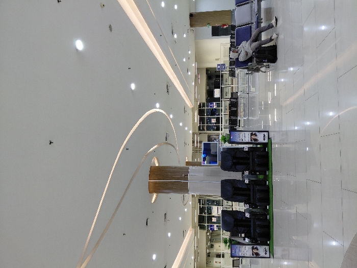 An airport waiting area shows rows of seats with a person resting on them. Foreground showcases a display of massage chairs lined up on a small green mat. Overhead are recessed lights and architectural arcs along the ceiling. The setting appears to be a modern and spacious interior of a transit hub.