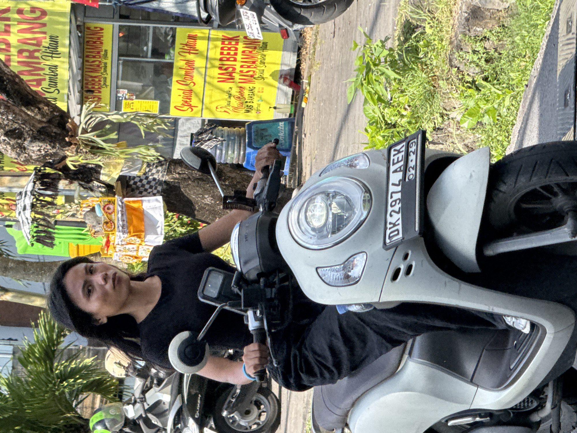 A person sits astride a light grey moped with the license plate "DK 2974 AEK", valid until February 2029. The person has long dark hair and wears a black t-shirt and a black beanie-style hat. The moped is parked on a street with other mopeds nearby. In the background, there are commercial signs advertising "Nasi Bebek Mas Bambang" and "Spesial Sambal Hitam," indicating the location is potentially near a restaurant or food vendor. The presence of Balinese-style black and white checkered cloth on a tree suggests the location is in Bali, specifically the city of North Kuta, Indonesia. Vegetation and a sidewalk are also visible.