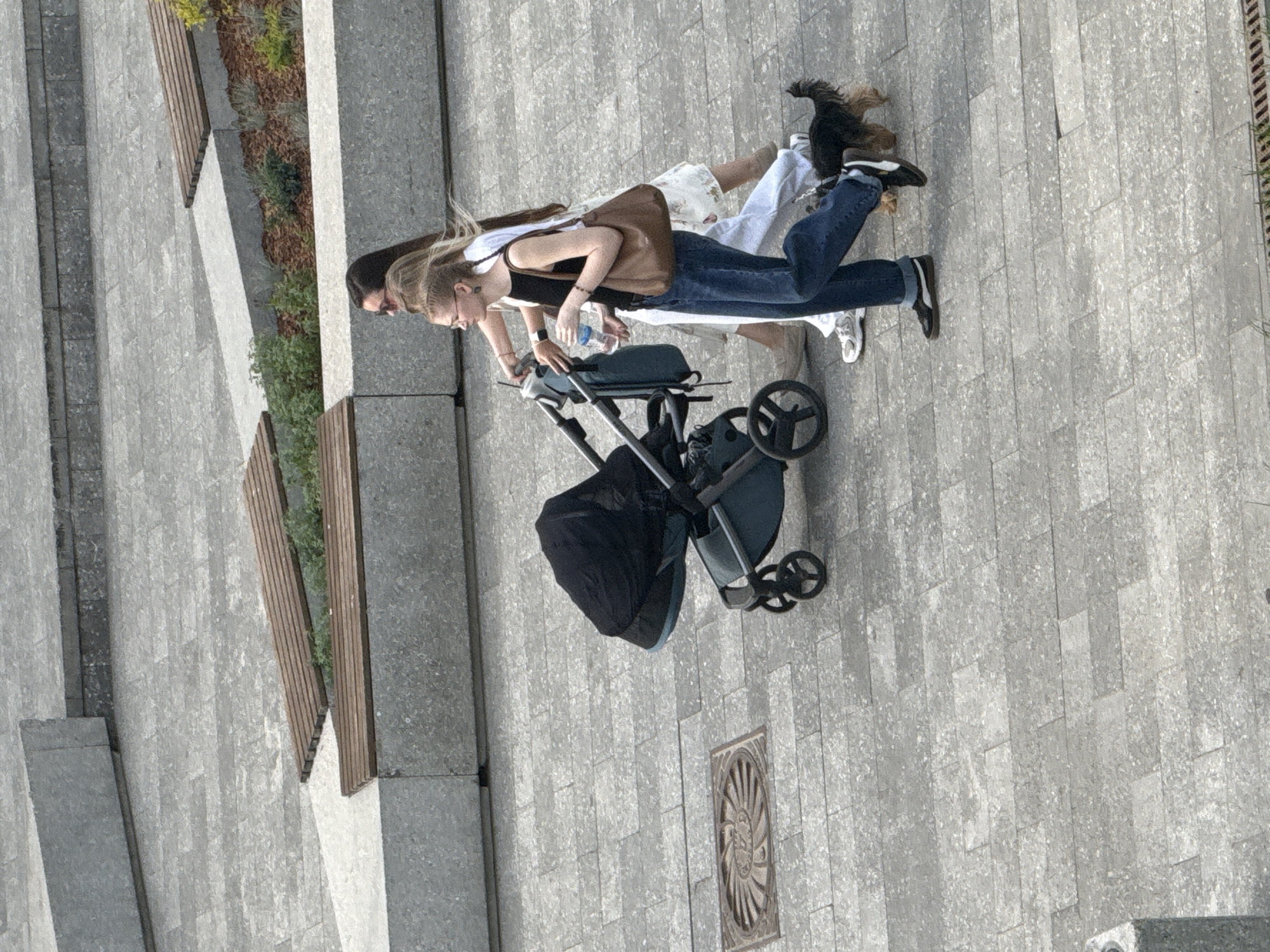 Two individuals walk along a paved walkway in Dmitrov, Russia, one pushing a black baby stroller. The individual pushing the stroller is wearing jeans, a black tank top, and a brown purse. The other person is wearing white pants and a white top, with a small dog in their arms. The paved walkway is constructed of rectangular stones. Adjacent to the walkway are concrete planters with wooden benches and sparse vegetation. A square metal grate is visible in the walkway.