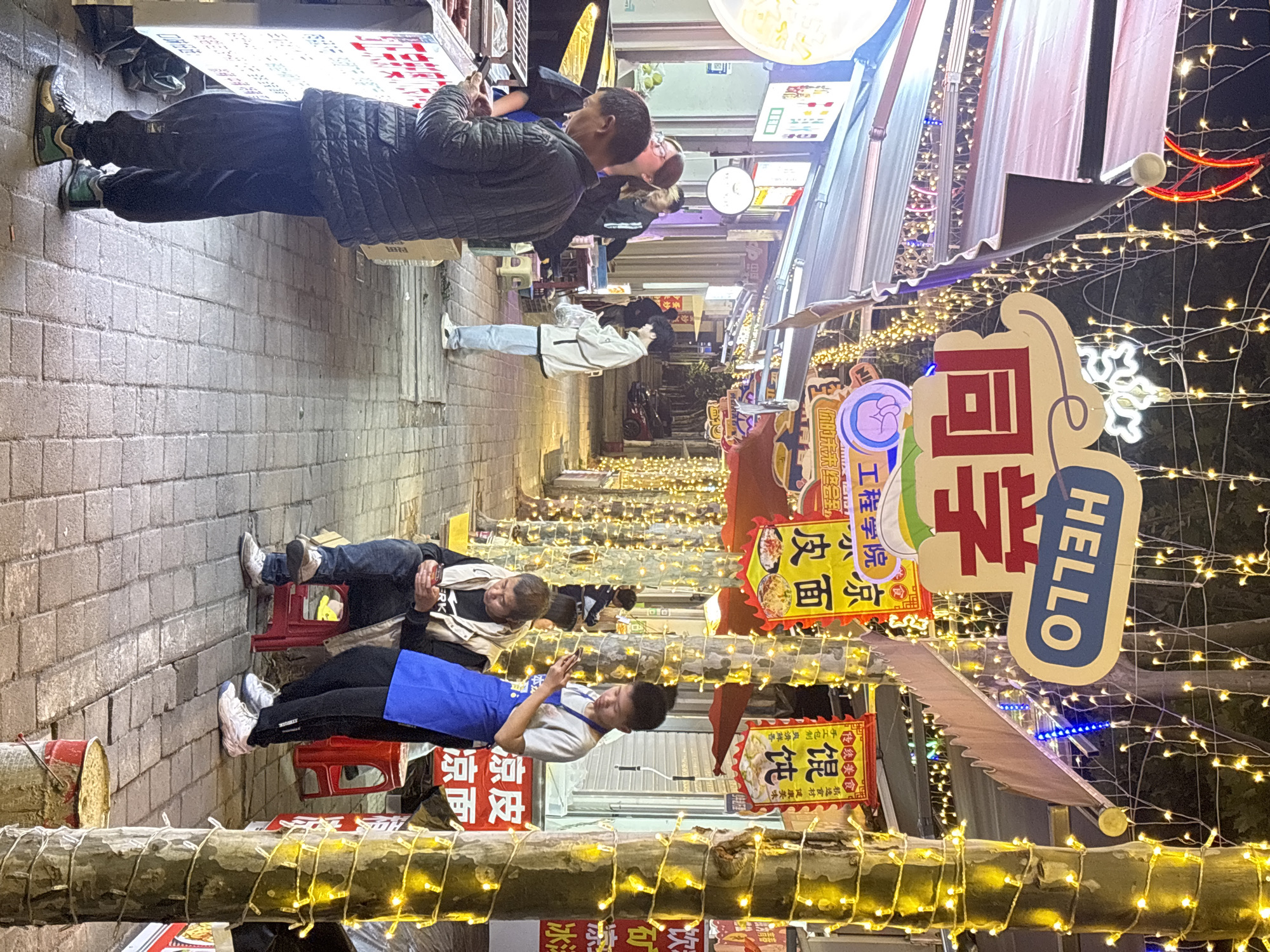 The image shows a street scene in the city of Jiangjin, China. The street is lined with food stalls, decorated with lights and signage, including signs with Chinese characters and the word "HELLO" in English. Several people are present; some are standing and looking at their phones, while others are seated on small stools. One person wears a blue apron. The street is paved with brick, and lights are strung overhead. The atmosphere is bustling and lively.