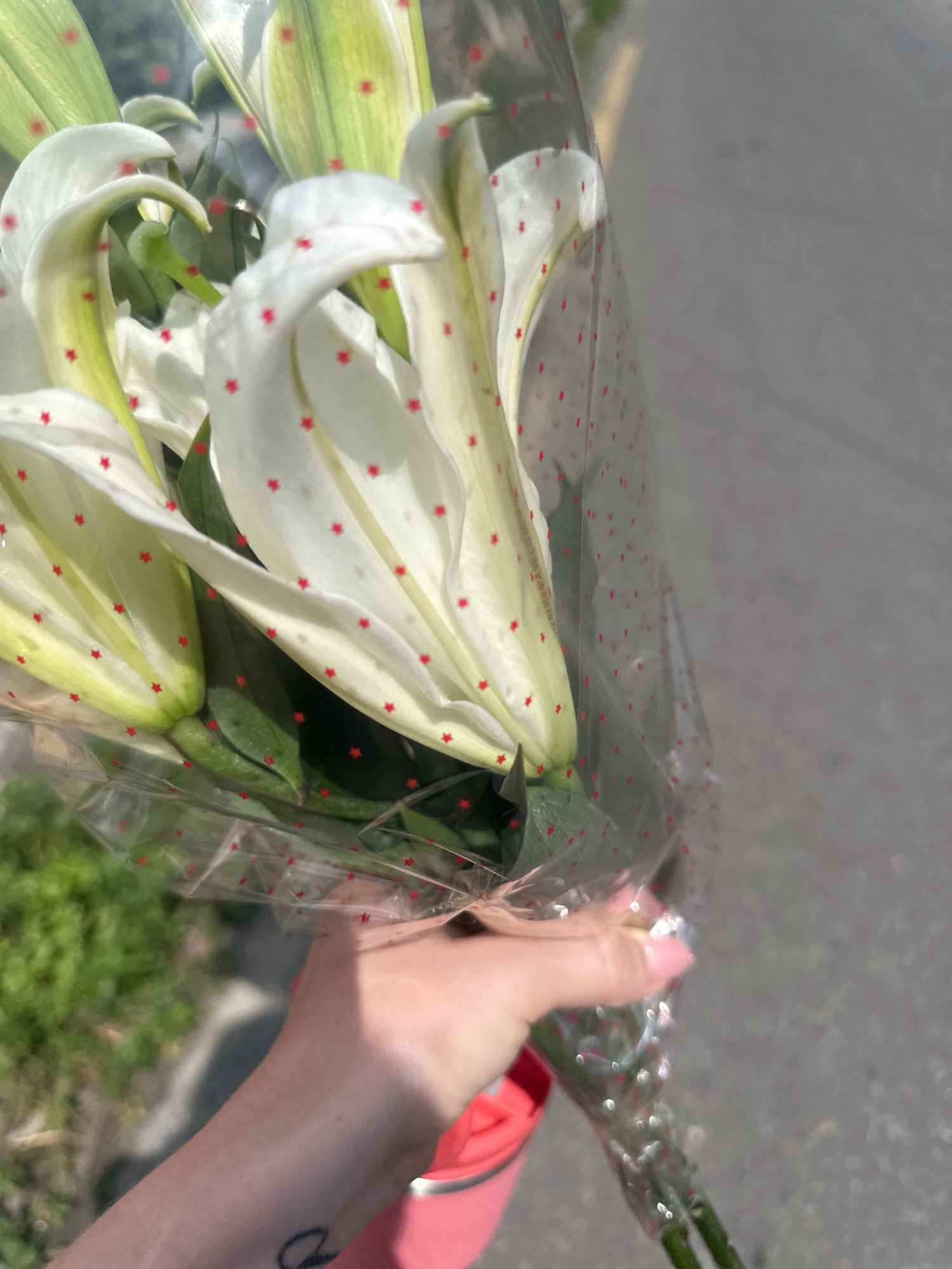 A close-up shot shows a person's hand holding a bouquet of white lilies. The lilies are partially wrapped in clear plastic adorned with small red star patterns. The background is blurred, revealing a gray paved surface, possibly a street, and some green foliage on the left. The lighting suggests it's daytime, with sunlight creating highlights on the plastic wrap and casting shadows. The scene appears to be outdoors in an urban environment. The specific location of Lalitpur, Nepal, is not discernable from the visual information provided in the image.