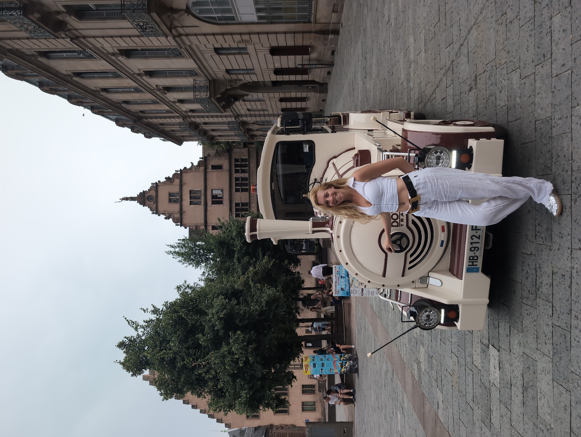 A woman with blonde curly hair, wearing a white crop top and white linen pants, stands next to a cream-colored tourist train in Strasbourg, France. The train has "DOUCH TRAIN" written on it. In the background, there are historic buildings with gabled roofs and balconies, and trees line the cobblestone street. The sky is overcast. The woman is smiling and appears to be posing for the photo. Several people are visible in the background, some sitting on steps and others walking. The scene suggests a pleasant day for sightseeing in the city.