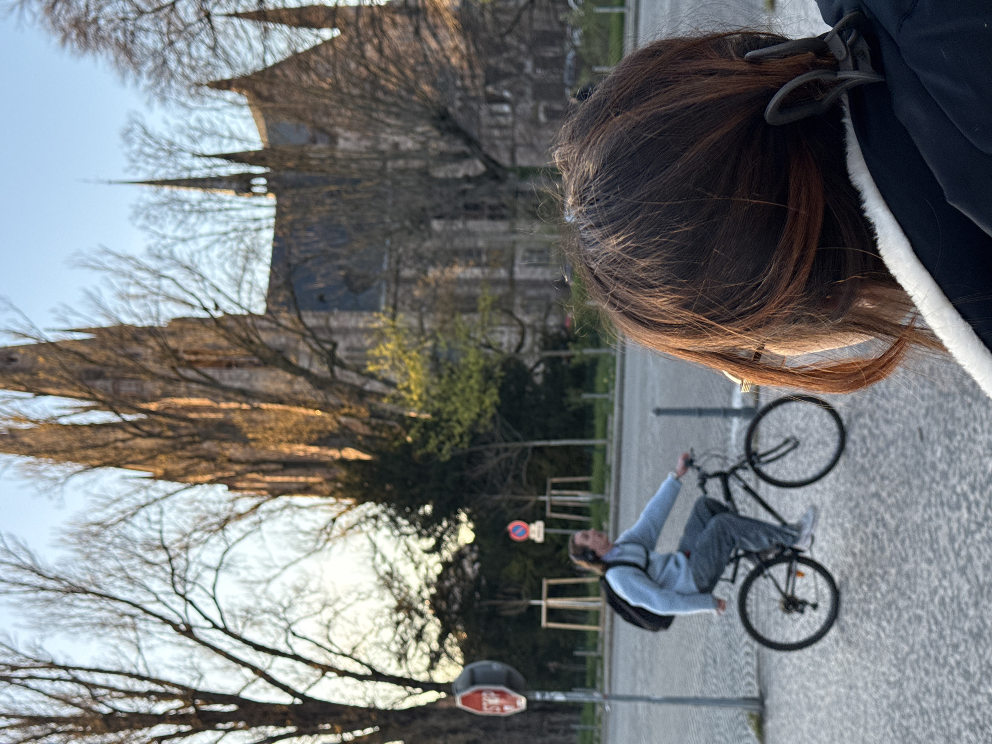 The image depicts a street scene in Strasbourg, France, with the Église Saint-Paul prominent in the background. The perspective is from behind and slightly to the right of a person with brown hair, tinged with red, secured by a dark hair clip, wearing a dark jacket with a white fur-lined collar. In the midground, a male individual is riding a black bicycle from left to right across a cobblestone street. He wears a light blue long-sleeved shirt, dark pants, white sneakers, a black backpack, and headphones. To the left, a red hexagonal STOP sign is visible on a pole, and further back, another round red and blue traffic sign. The background features the distinctive neo-Gothic architecture of the Église Saint-Paul, characterized by multiple spires and dark stone, partially obscured by numerous bare deciduous trees, some with hints of new green foliage. The sky is clear and light blue, and the scene is illuminated by warm, low-angle sunlight, indicating either dawn or dusk.