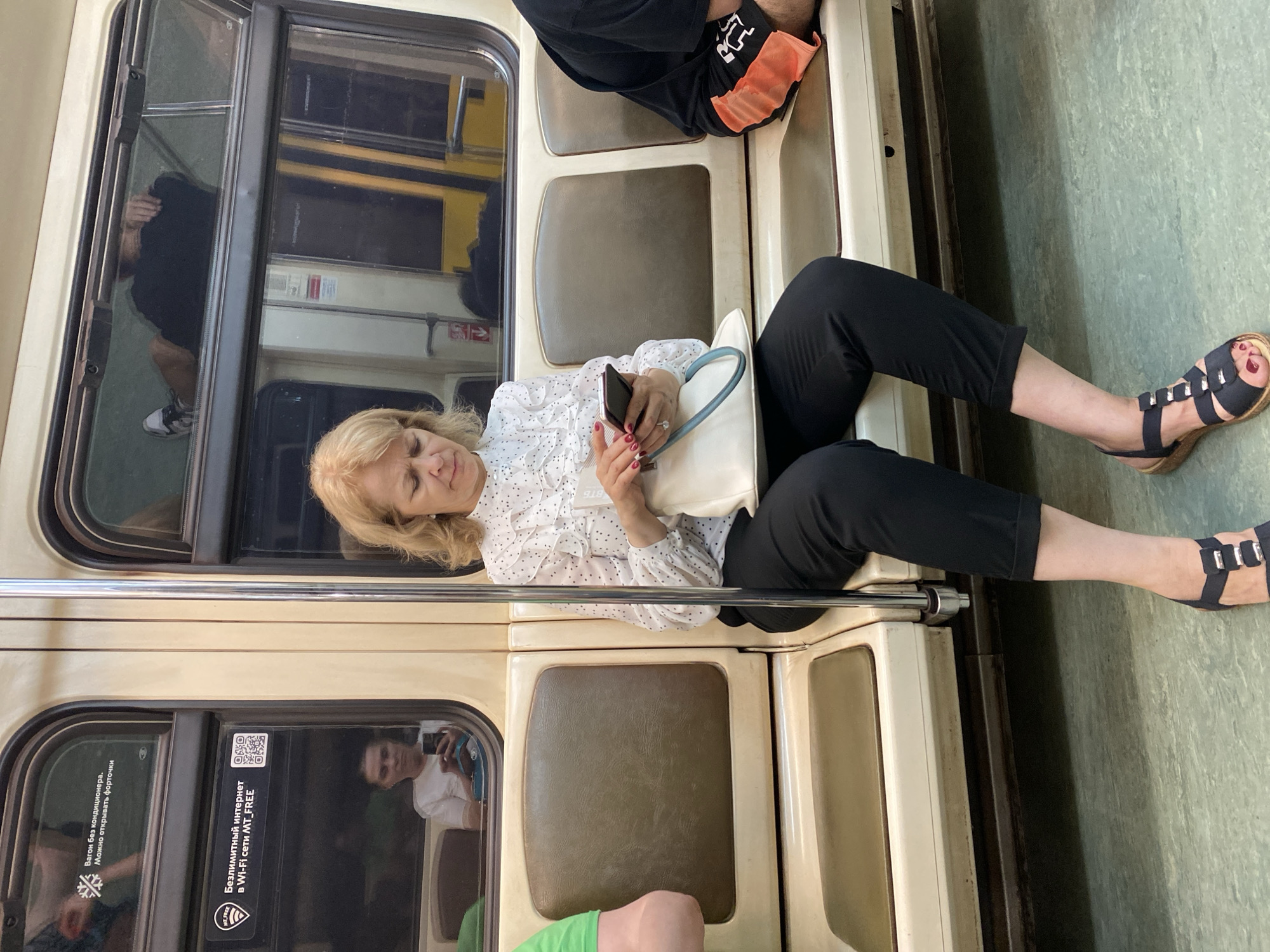 A woman is seated on a train. She has blonde hair, a white polka dot shirt, and black capri pants. She is wearing black sandals with a block heel. She is holding a cell phone in her hands and looking at it. She has a white purse. There is a man seated next to her, and a man behind her. The train car looks old and beige. The windows have stickers that say the train car has no air conditioning, and that there is free wifi. The train appears to be in Moscow, Russia.
