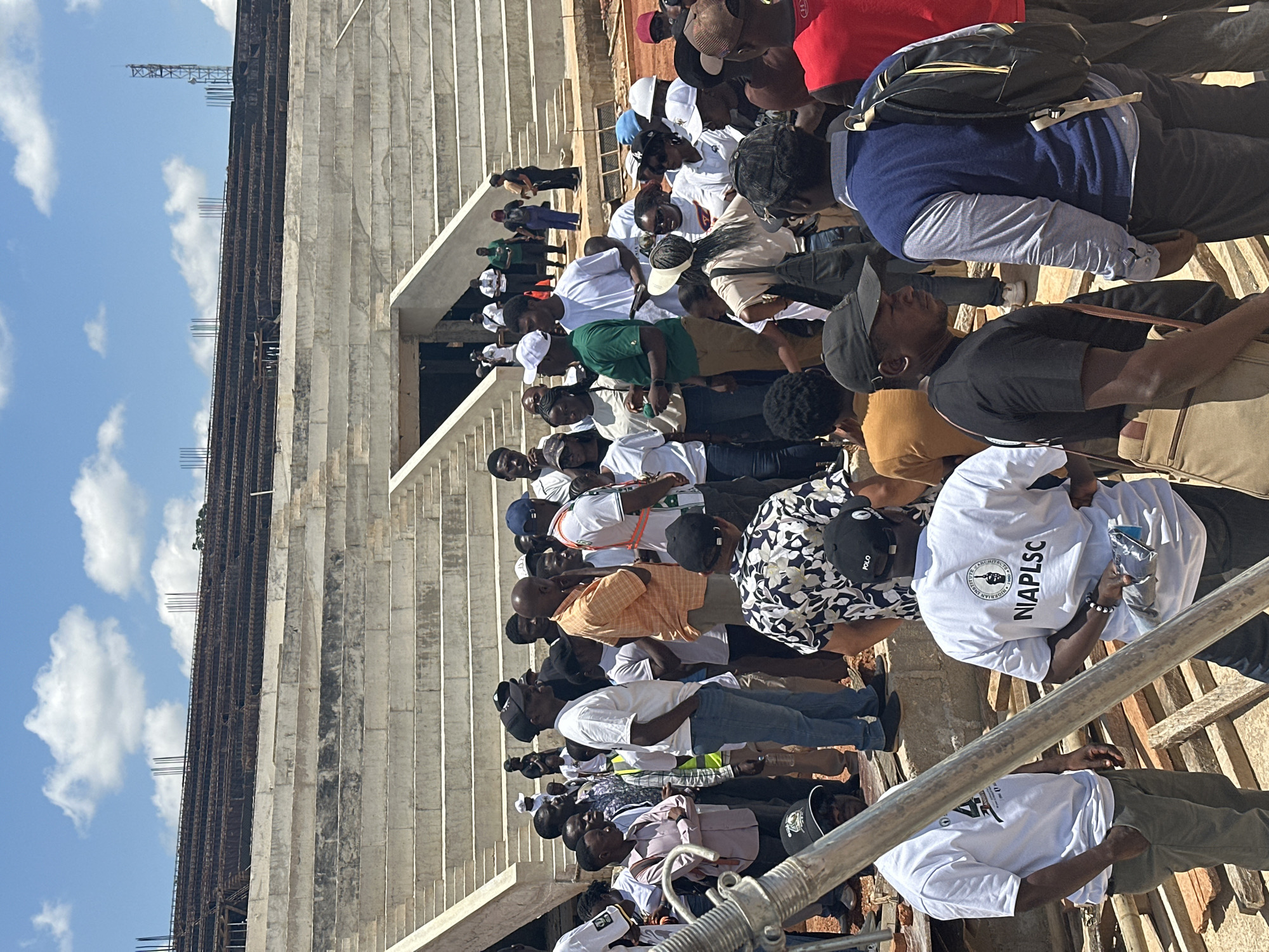 The image shows a gathering of people in front of a stadium under construction. The stadium has concrete seating tiers and exposed rebar at the top. The people are diverse in age and gender, dressed in casual attire. Some are wearing white t-shirts with logos, suggesting a group affiliation. The environment appears to be outdoors, with a blue sky and scattered clouds. The ground is uneven and appears to be a construction site, with some wooden planks visible. A man in the foreground is wearing a blue vest over a light blue shirt and a backpack. Another man is wearing a black t-shirt and a beige messenger bag. The people are standing and talking, with some appearing to be observing the construction. The location is specified as the city of Jos, Nigeria.
